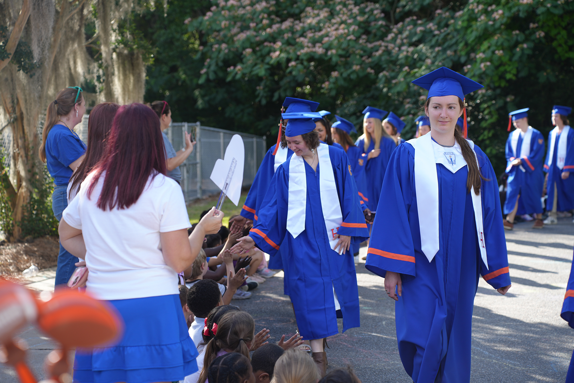 Graduates in blue gowns and hats walk past a crowd of children cheering. Trees and greenery in the background.