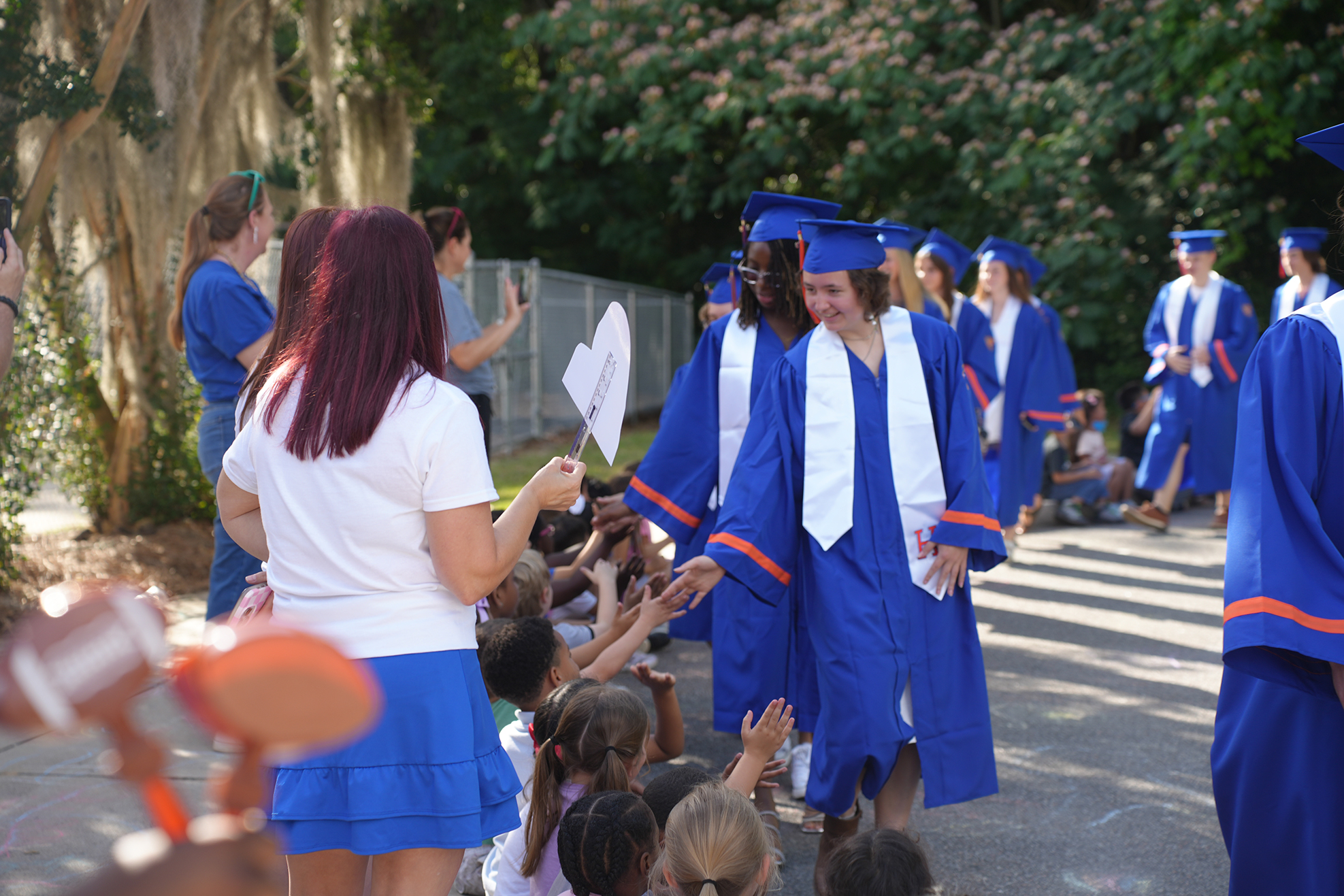 A group of graduates in blue gowns walk past a crowd, with children reaching out to touch them.