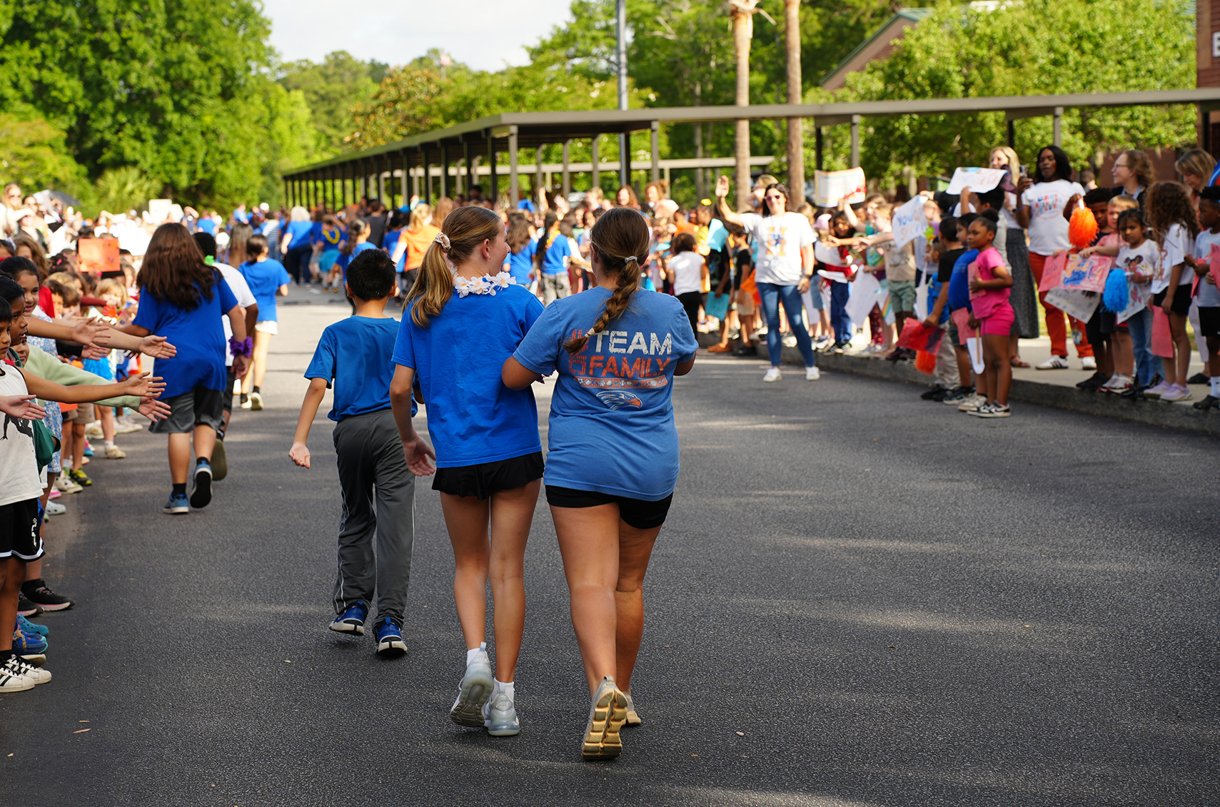 Several children and adults in blue shirts walk down a road with onlookers on either side.