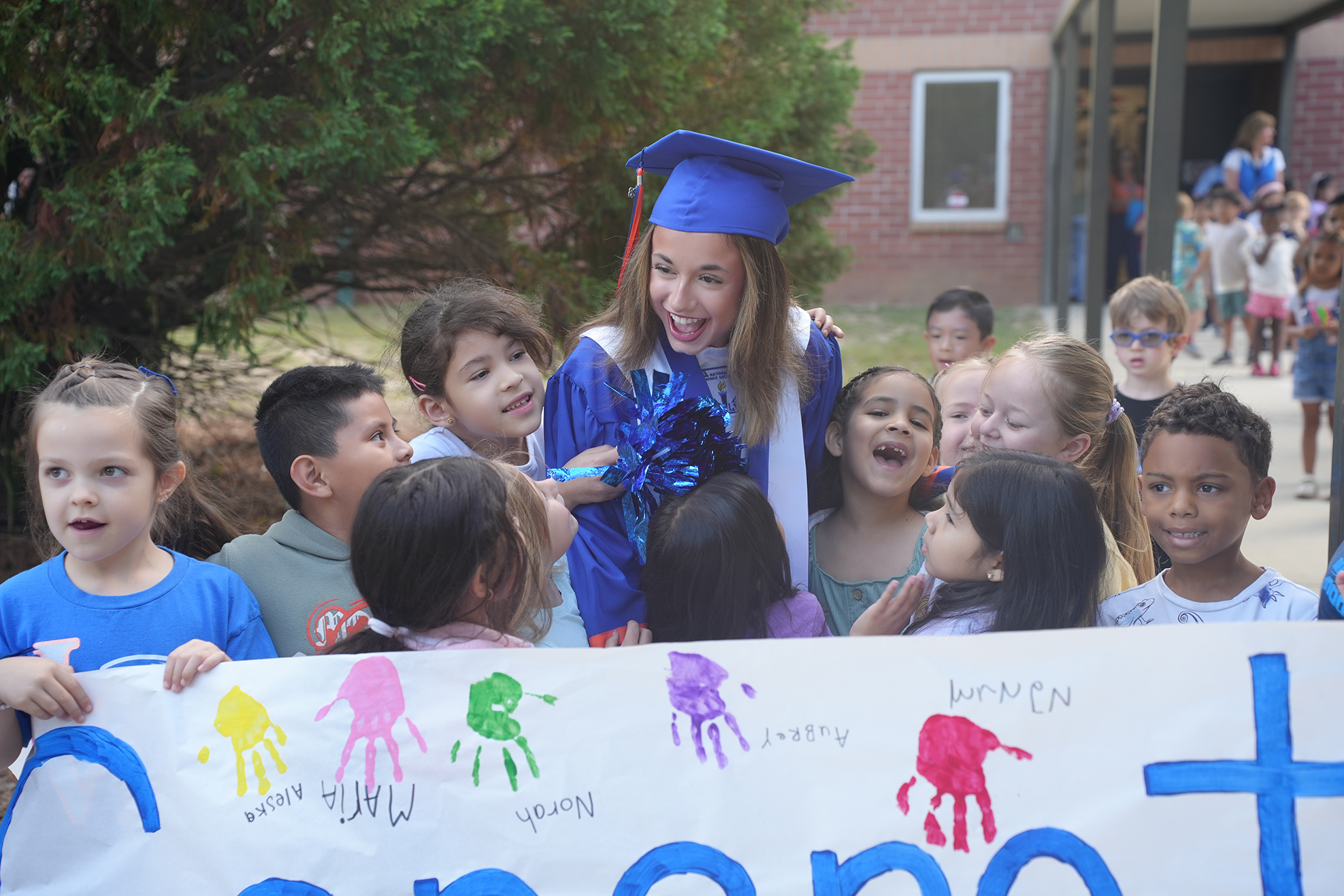 A group of young children holding a banner with painted hands, and a smiling young woman in a graduation gown.