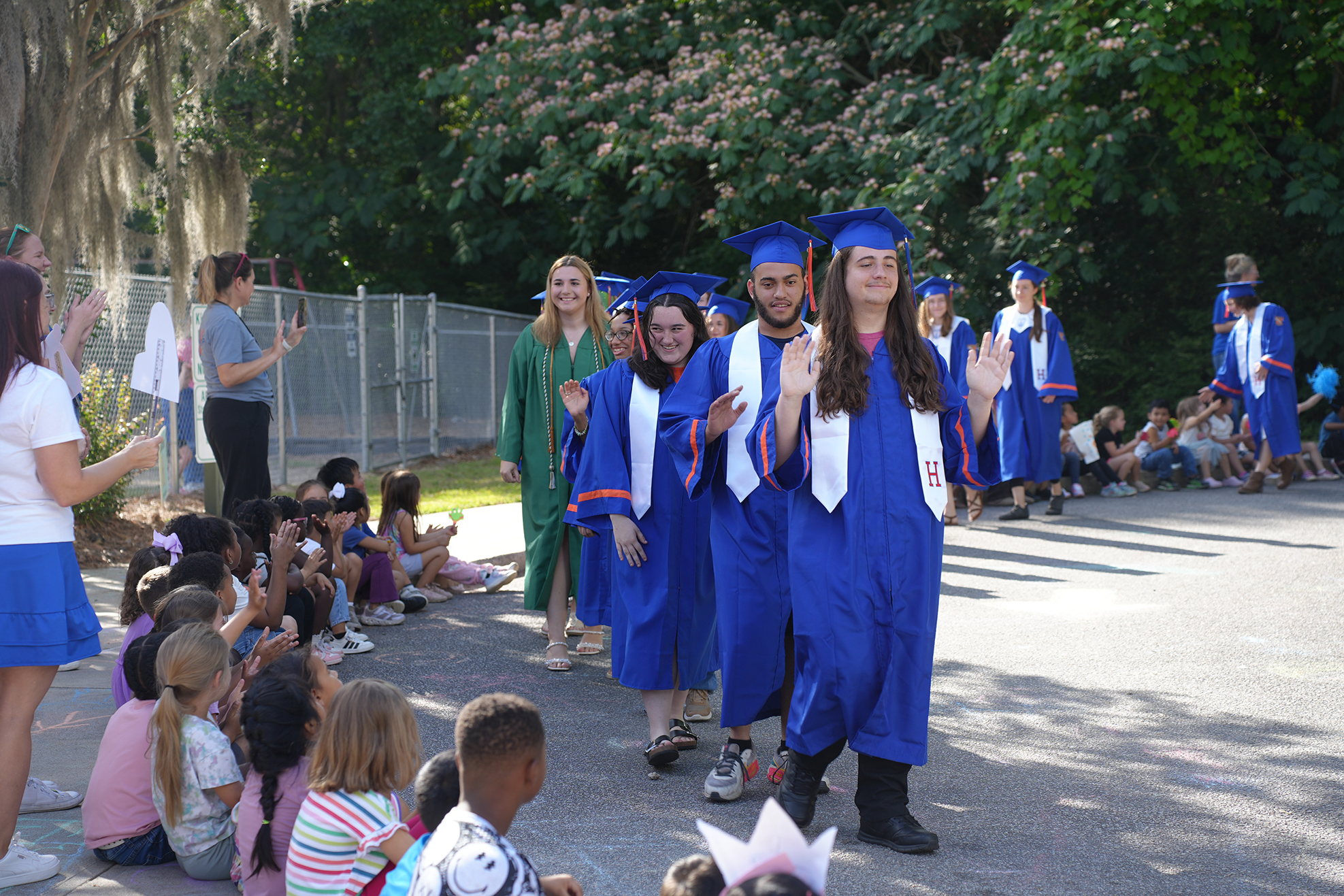 A group of people in blue gowns and hats walk by a crowd of seated children.