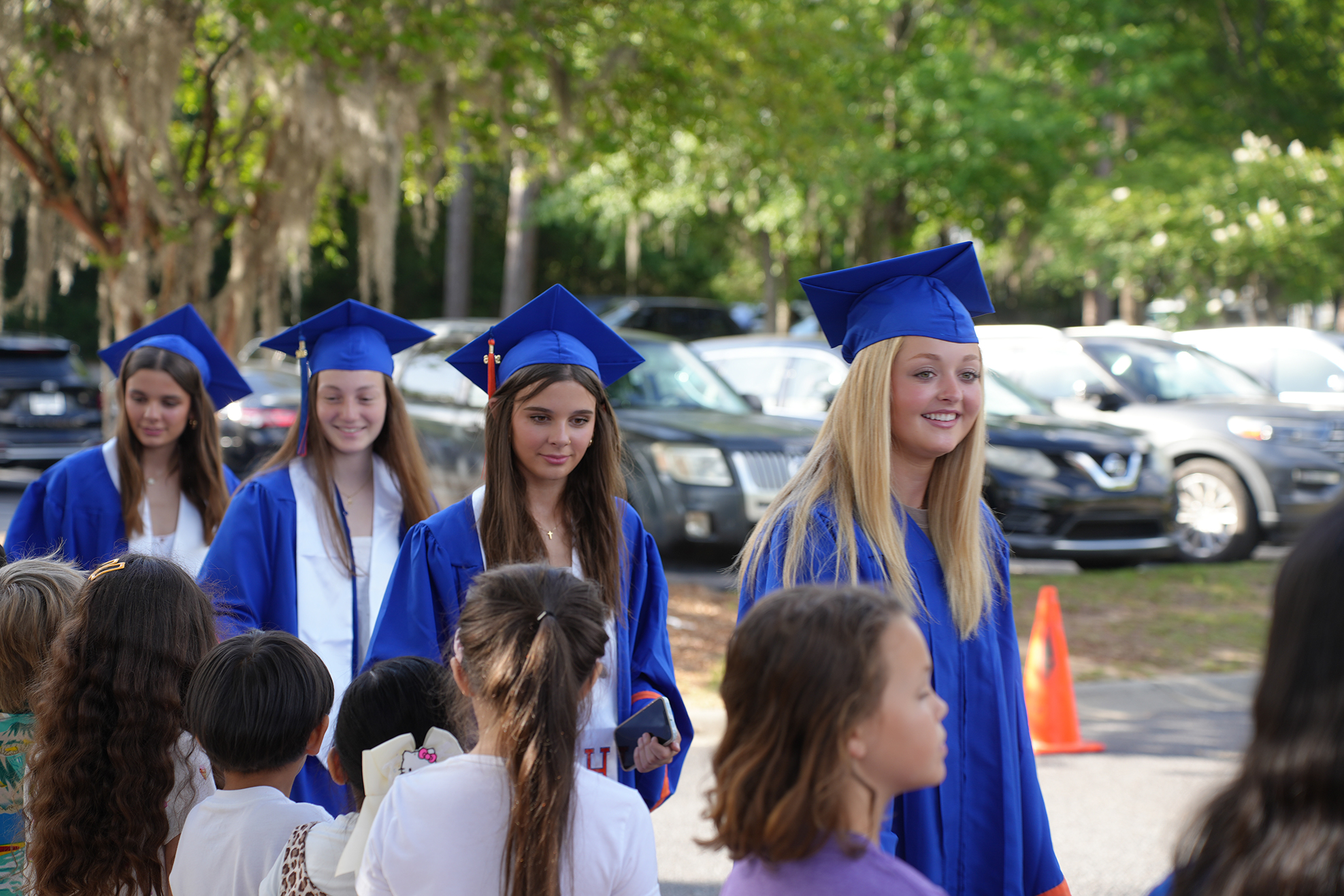 A group of women in blue graduation gowns and hats stand outside, smiling. Cars are parked behind them. A traffic cone is on the right.