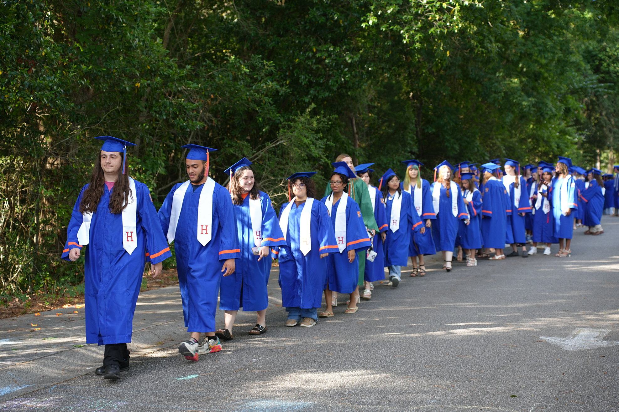 Many people in blue graduation gowns walk in a line on a road surrounded by green trees.