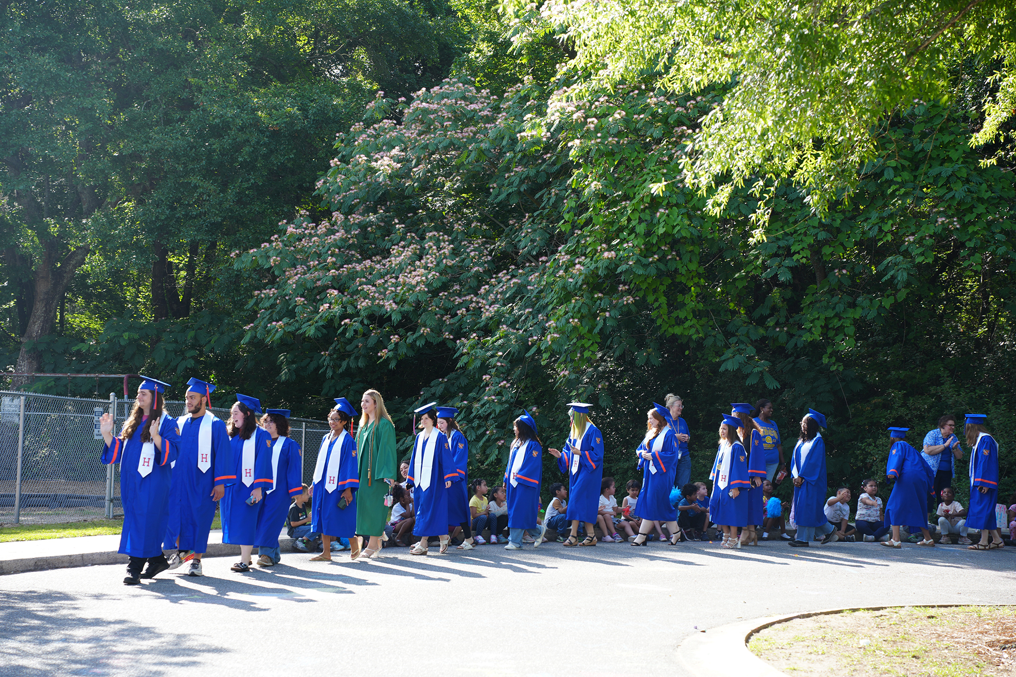 Graduates in blue gowns and caps stand in a row, surrounded by trees. Spectators stand behind them.