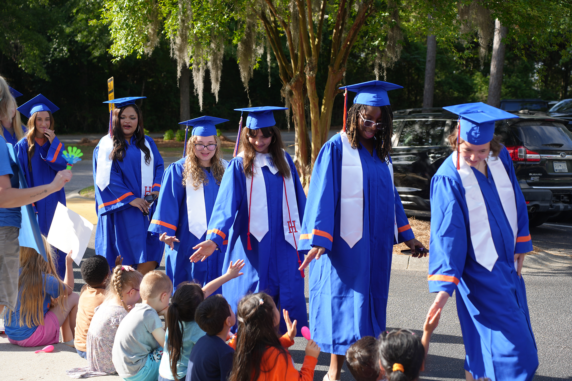 A group of individuals in blue graduation gowns and caps walking down a street, greeted by children.