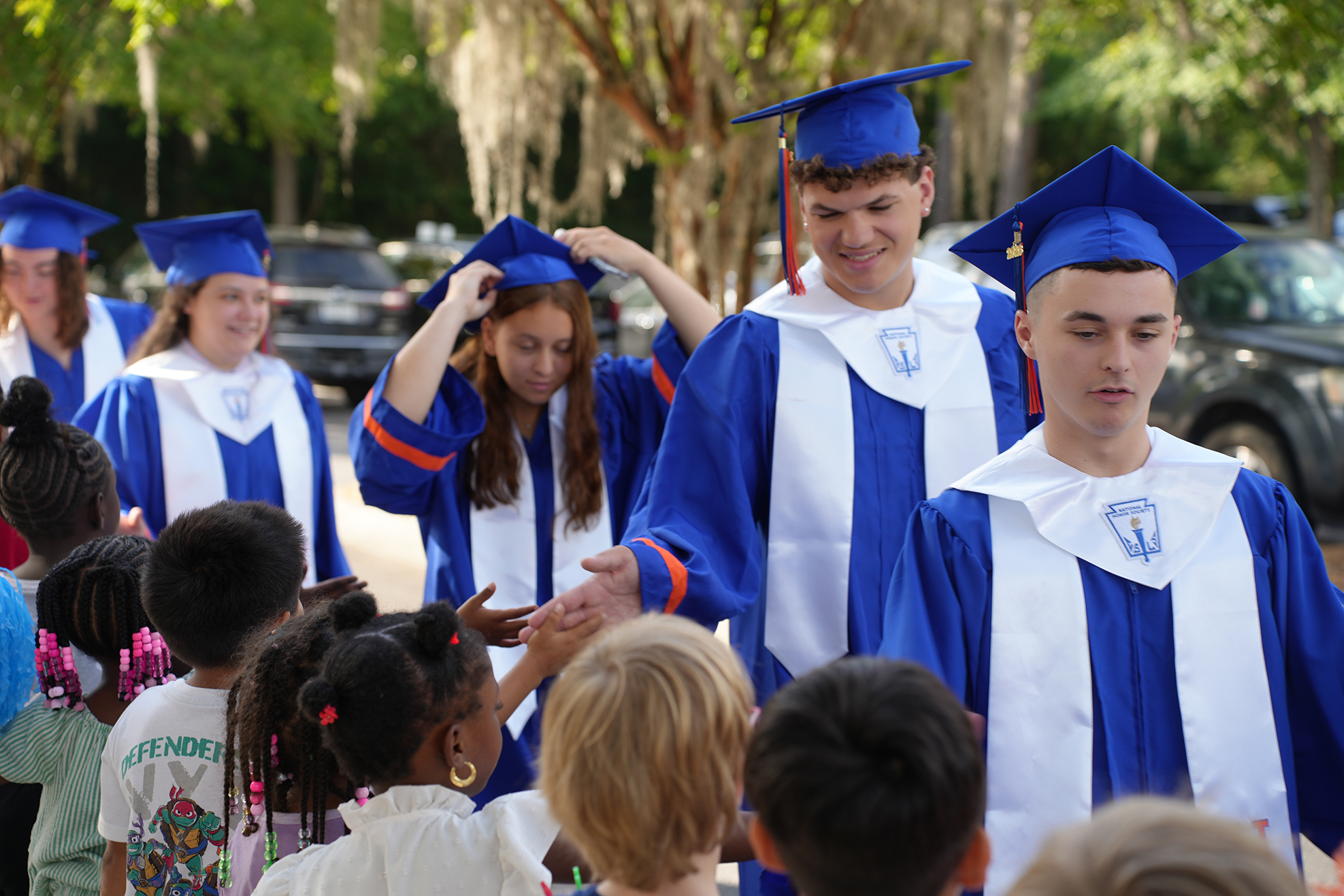 Graduates in blue and white caps and gowns interact with children in an outdoor setting.
