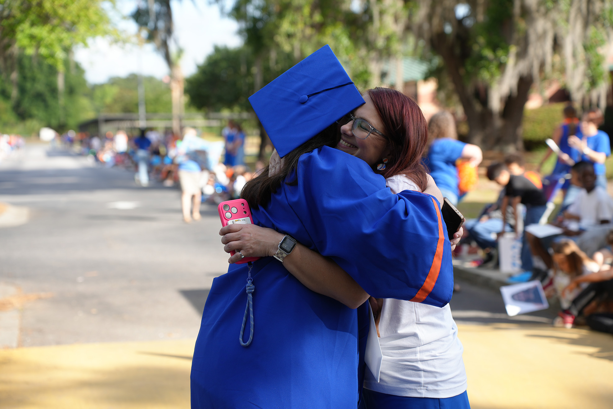 Two women hug outdoors in front of a road; one wears a blue gown, the other a white shirt.