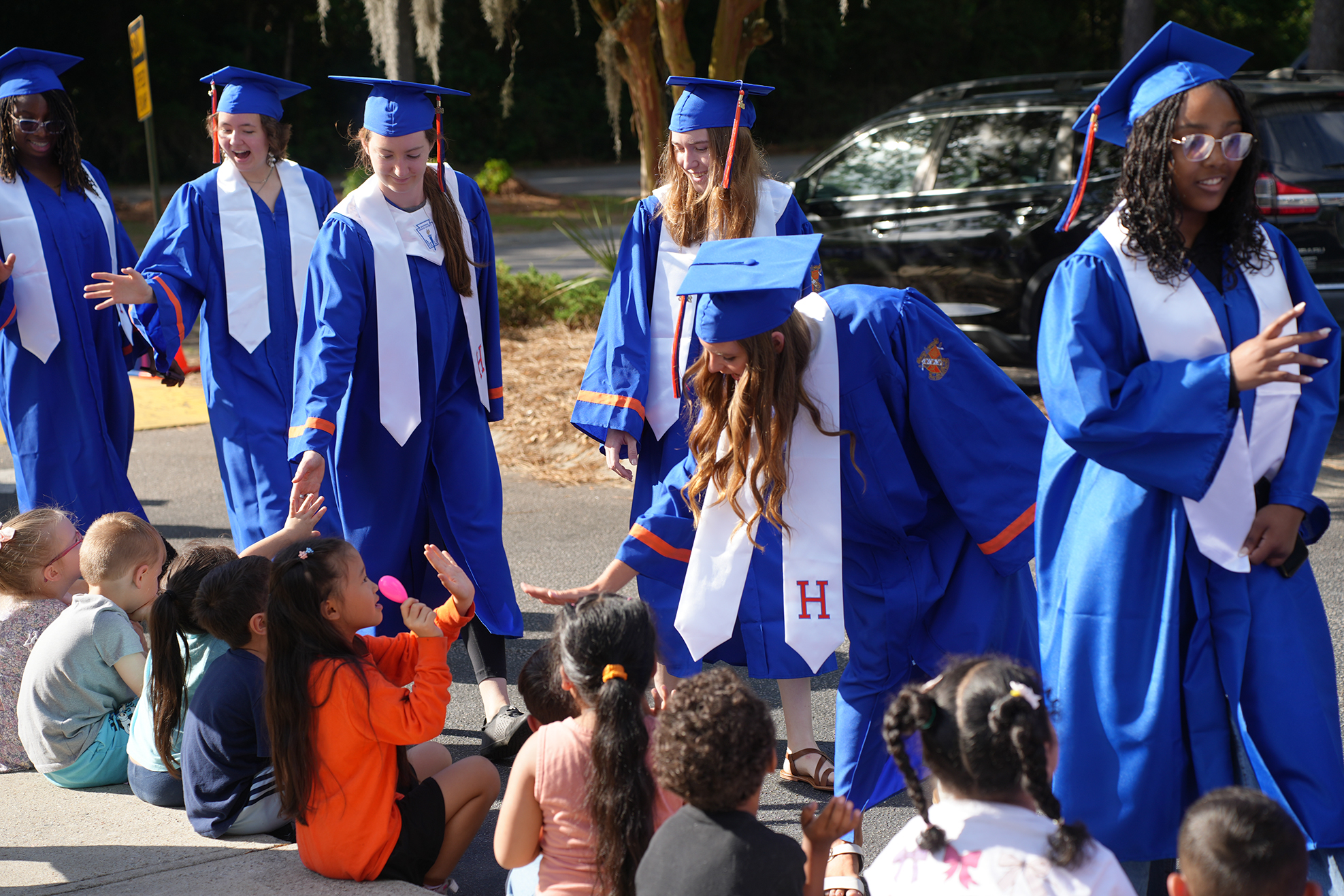 Several people in blue graduation gowns and hats, possibly teachers, talk to a group of children.