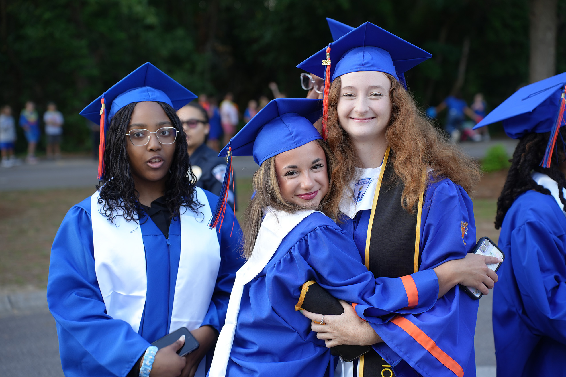 Four girls in blue and white graduation gowns and caps stand together. One girl hugs another from behind.