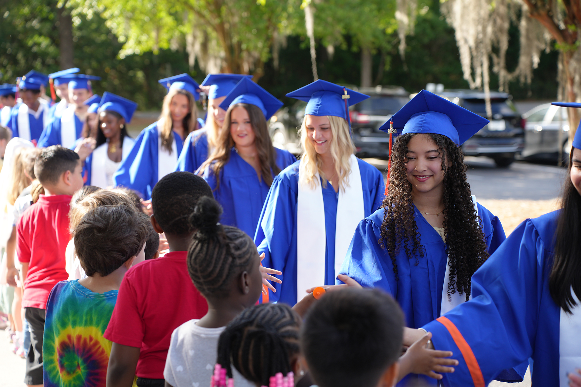 A group of students in blue graduation gowns and caps walk with children in a parking lot.