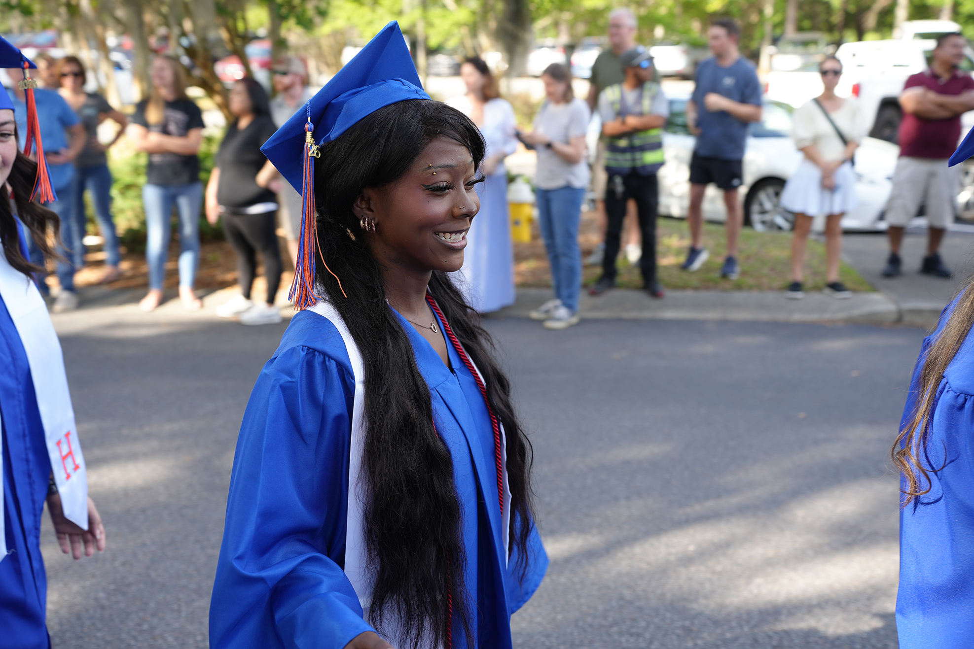 A woman with a blue gown and cap walks outdoors with others. Several people stand behind her.