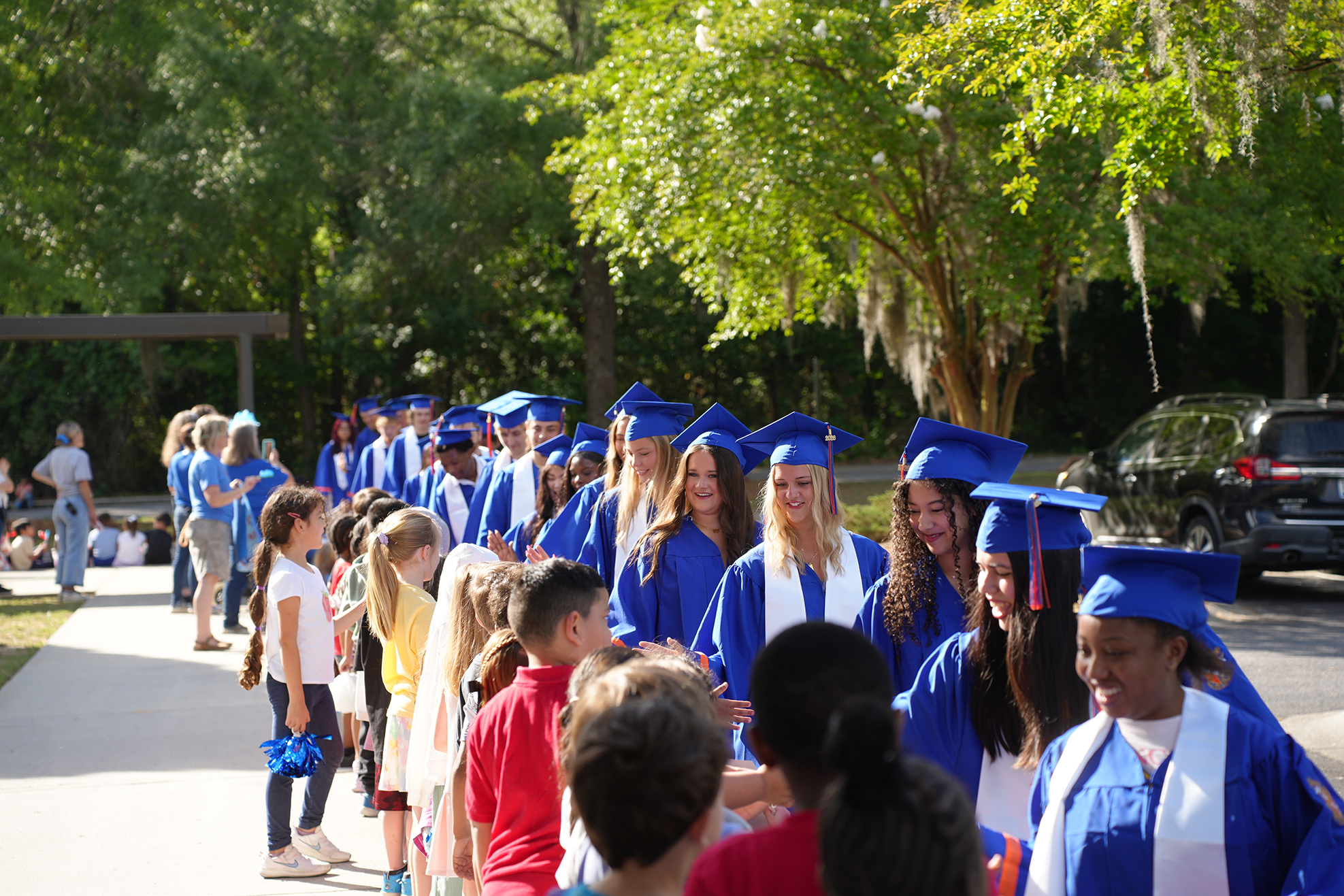 Graduates in blue gowns and hats line up and greet others in front of a car. Trees and a walkway are in the background.