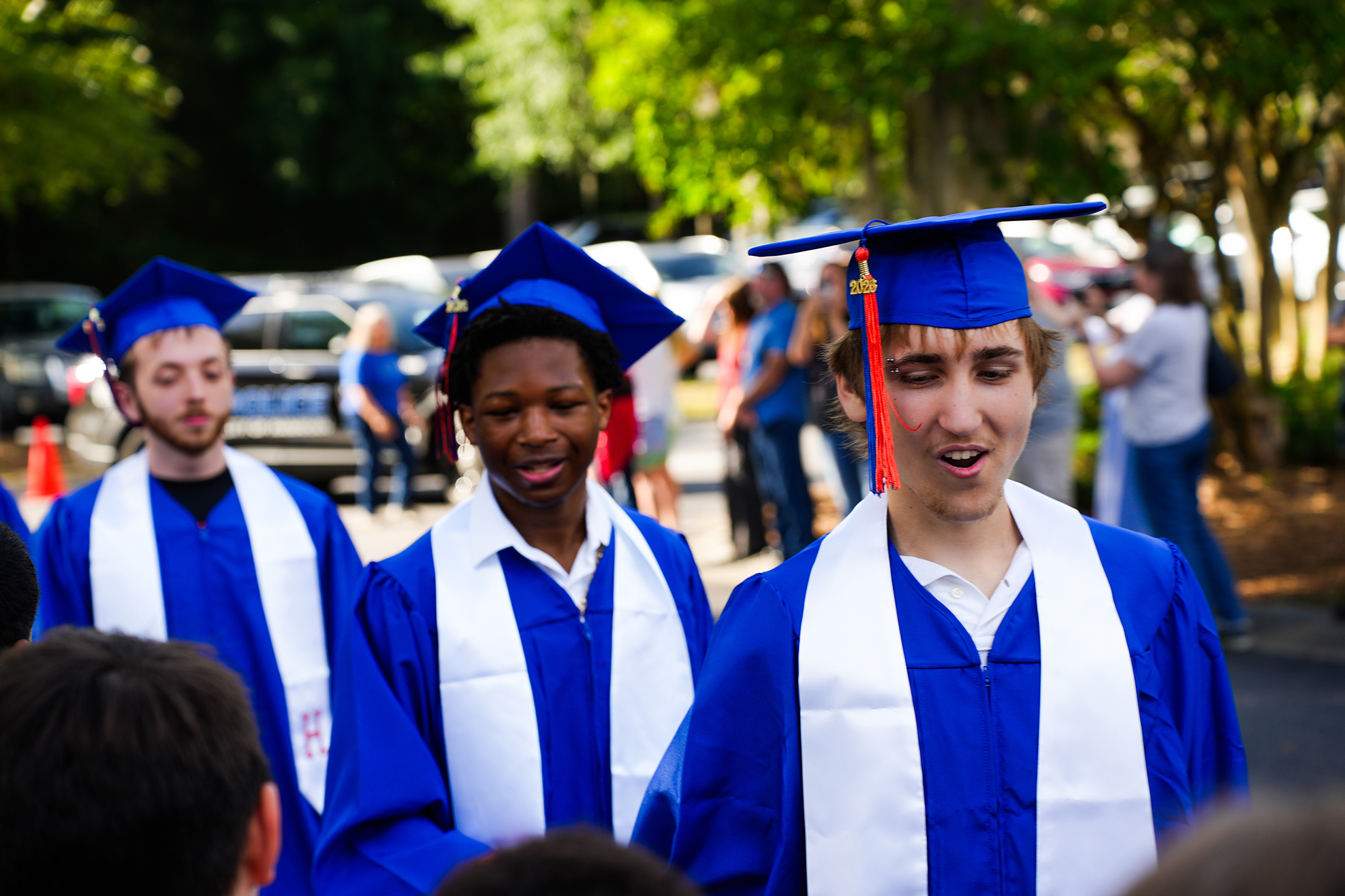 Three young people in blue caps and gowns are standing in an outdoor area, with a group of people walking behind them.