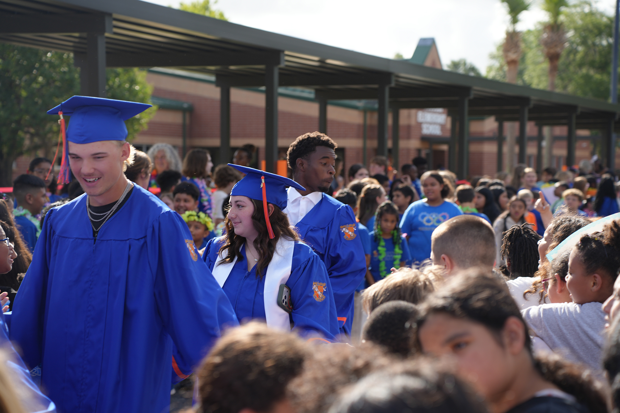 A crowd of students in blue gowns and caps walks down a hallway at a school.