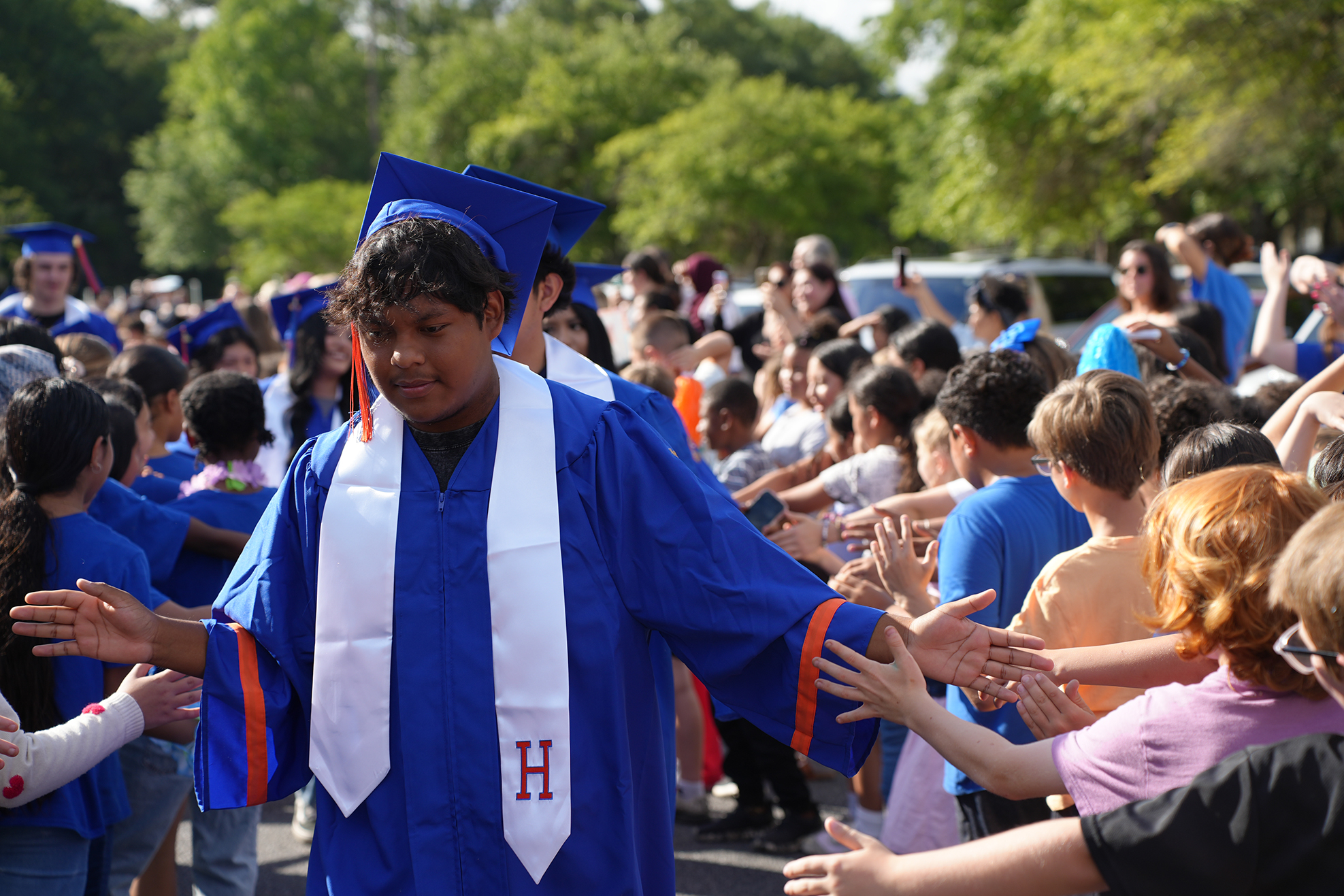 A person in a blue and white graduation gown stands in a crowd. The crowd appears to be cheering.