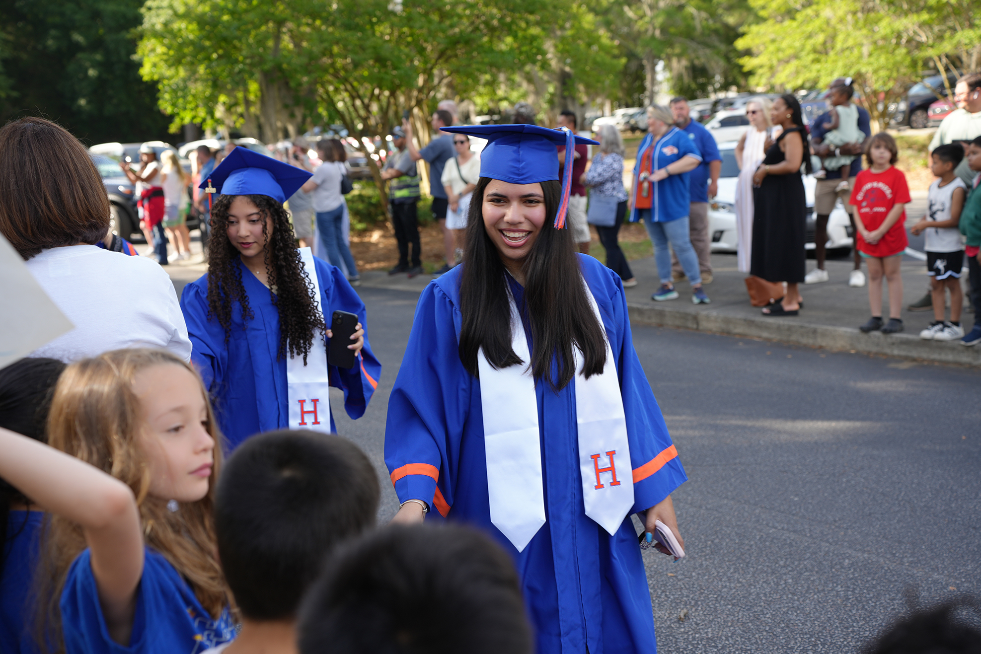 A woman in a blue graduation gown walks down a street, greeted by children in blue.