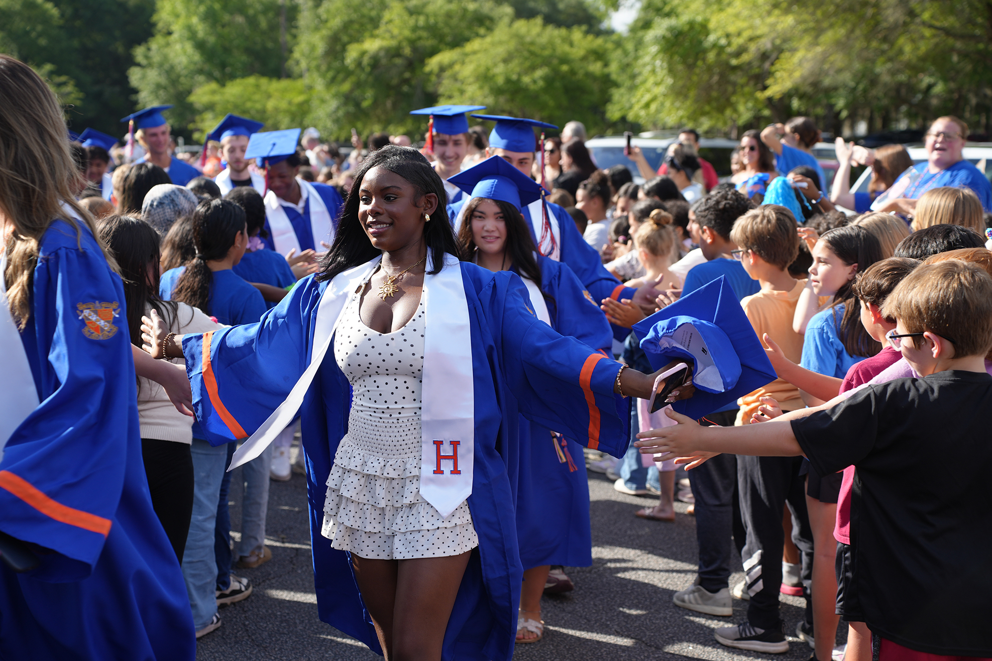 Graduates in blue caps and gowns walk through a crowd outdoors; some hold blue caps.