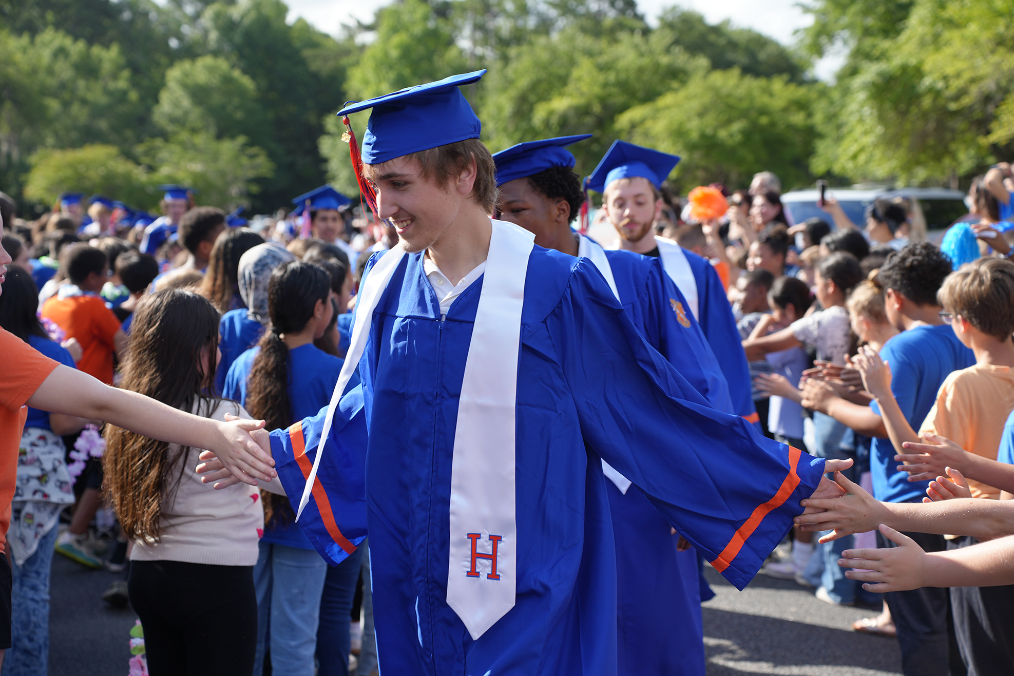 Graduates in blue gowns and hats, some shaking hands with others, outdoors. Trees and a crowd in background.