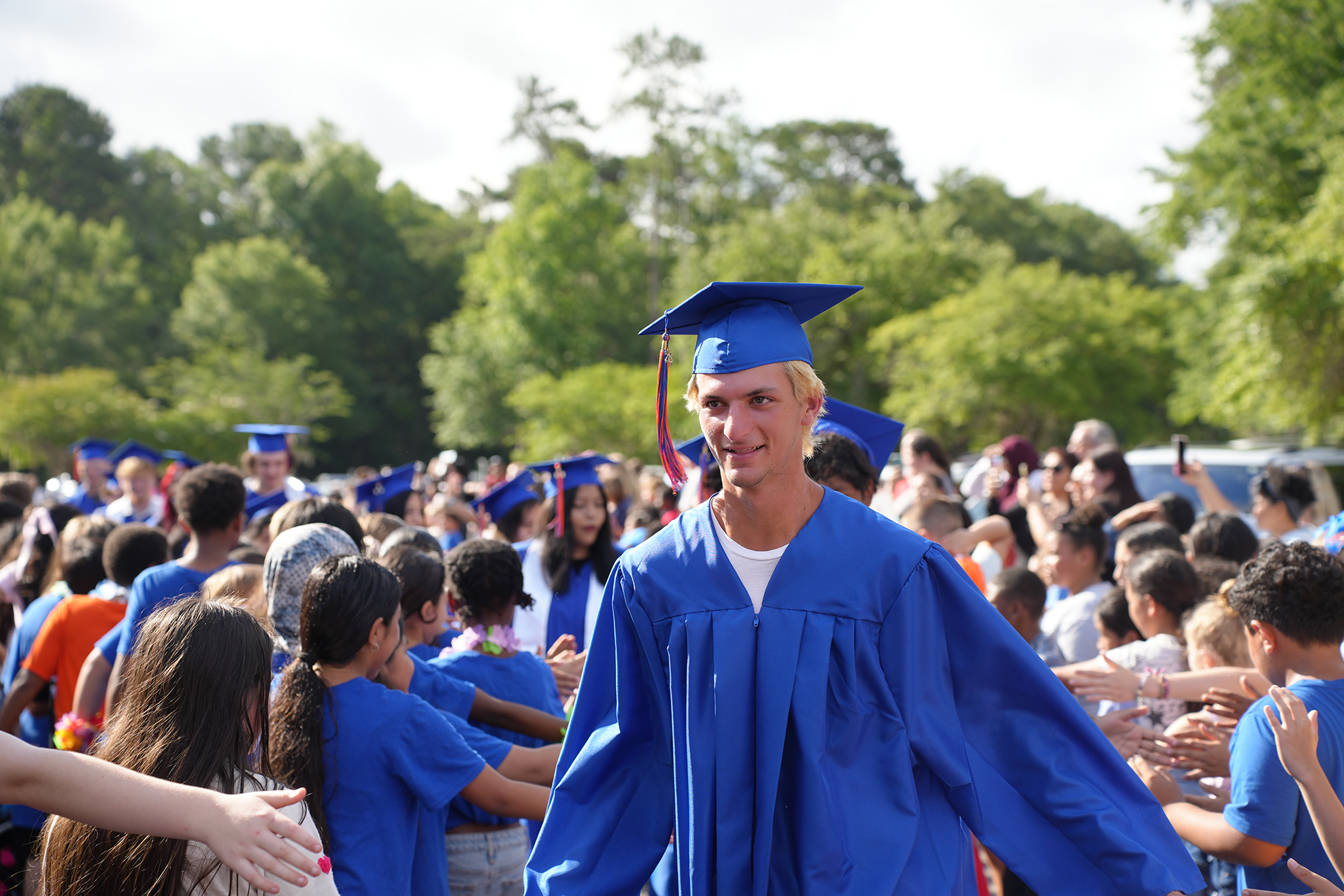 A person in a blue graduation gown walks through a crowd of people at a graduation ceremony.