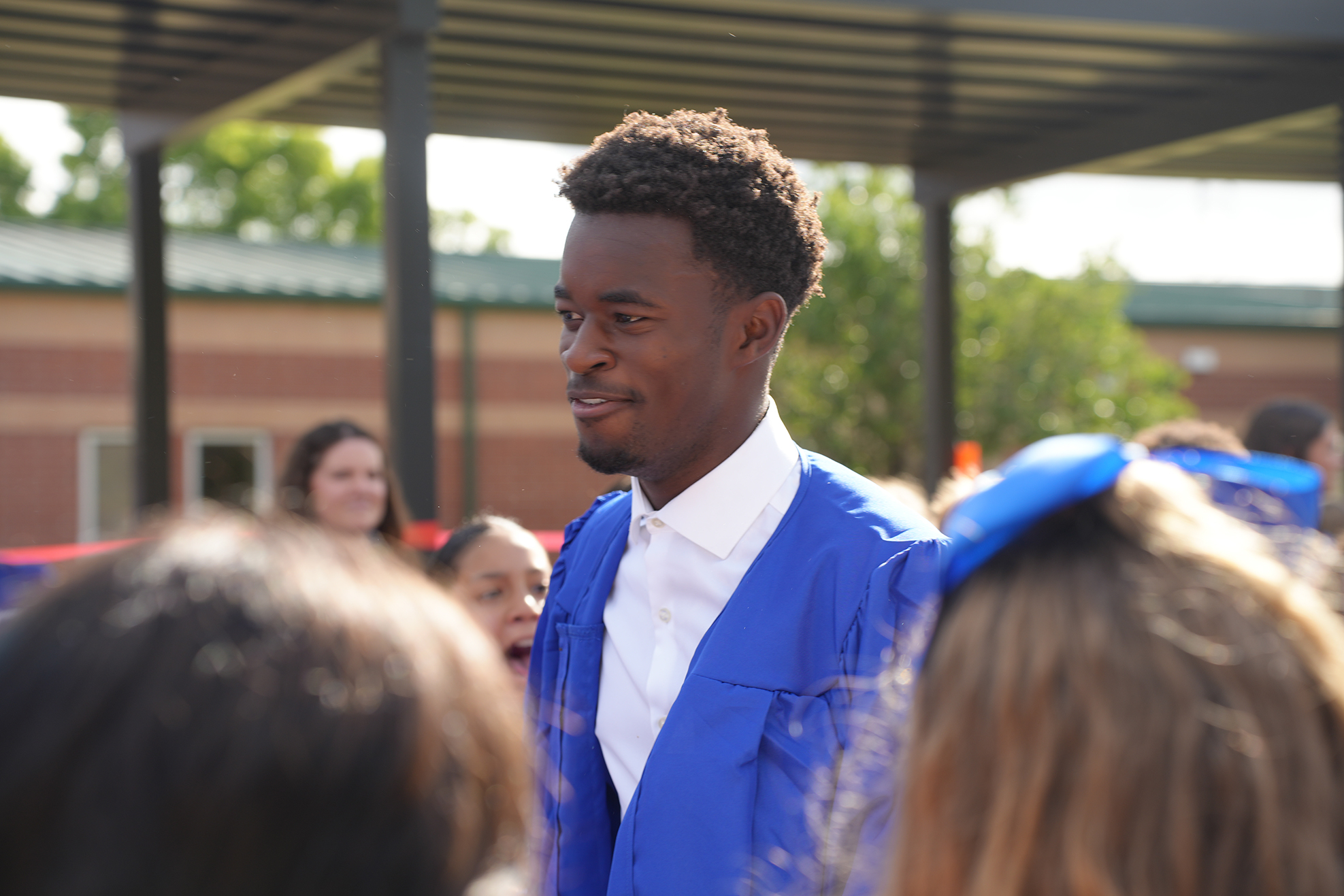 Man in blue cap and gown surrounded by others, one wearing blue bow, outside school.