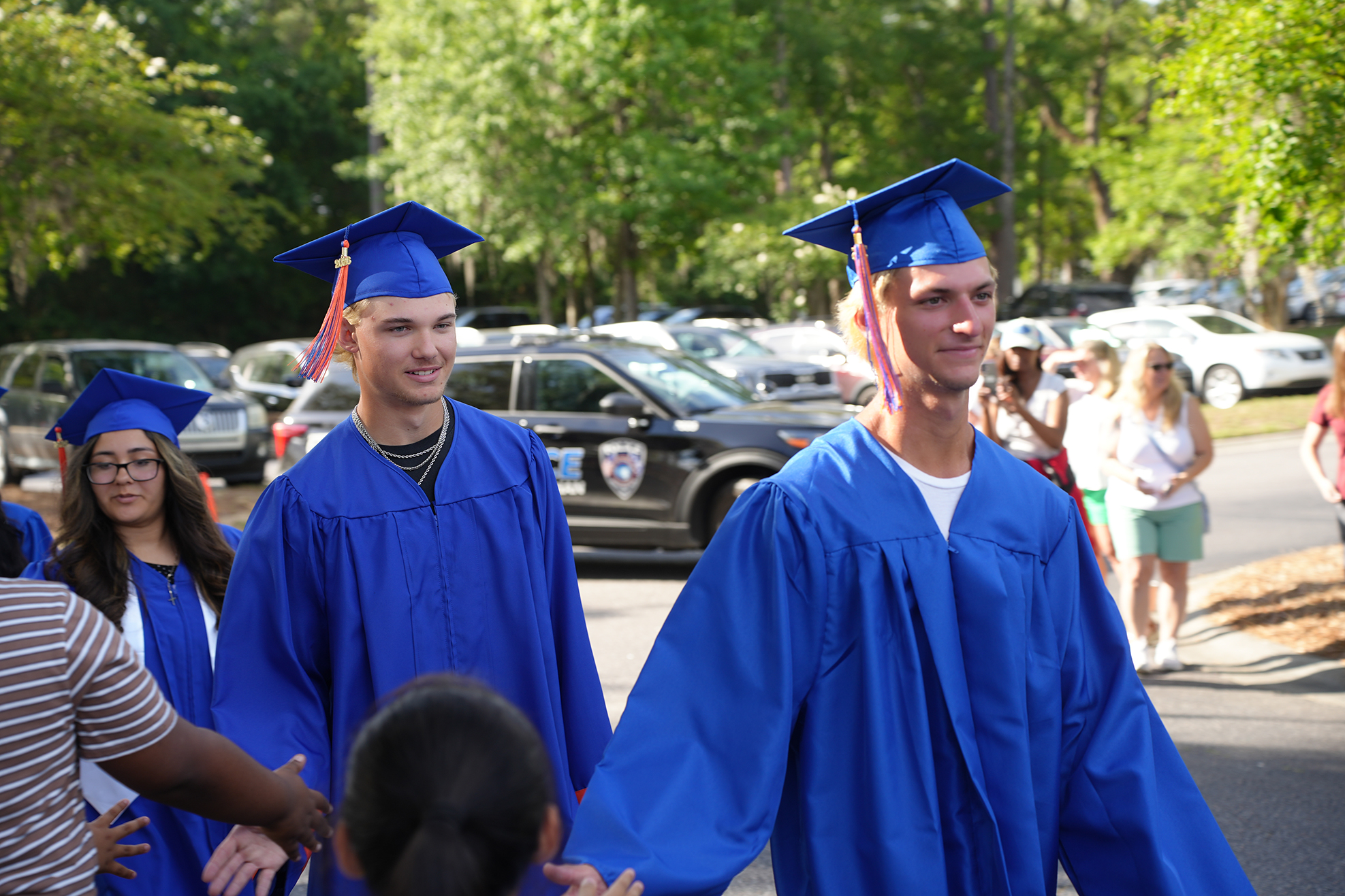 Two men in caps and gowns walk together in a parking lot with trees and parked cars.