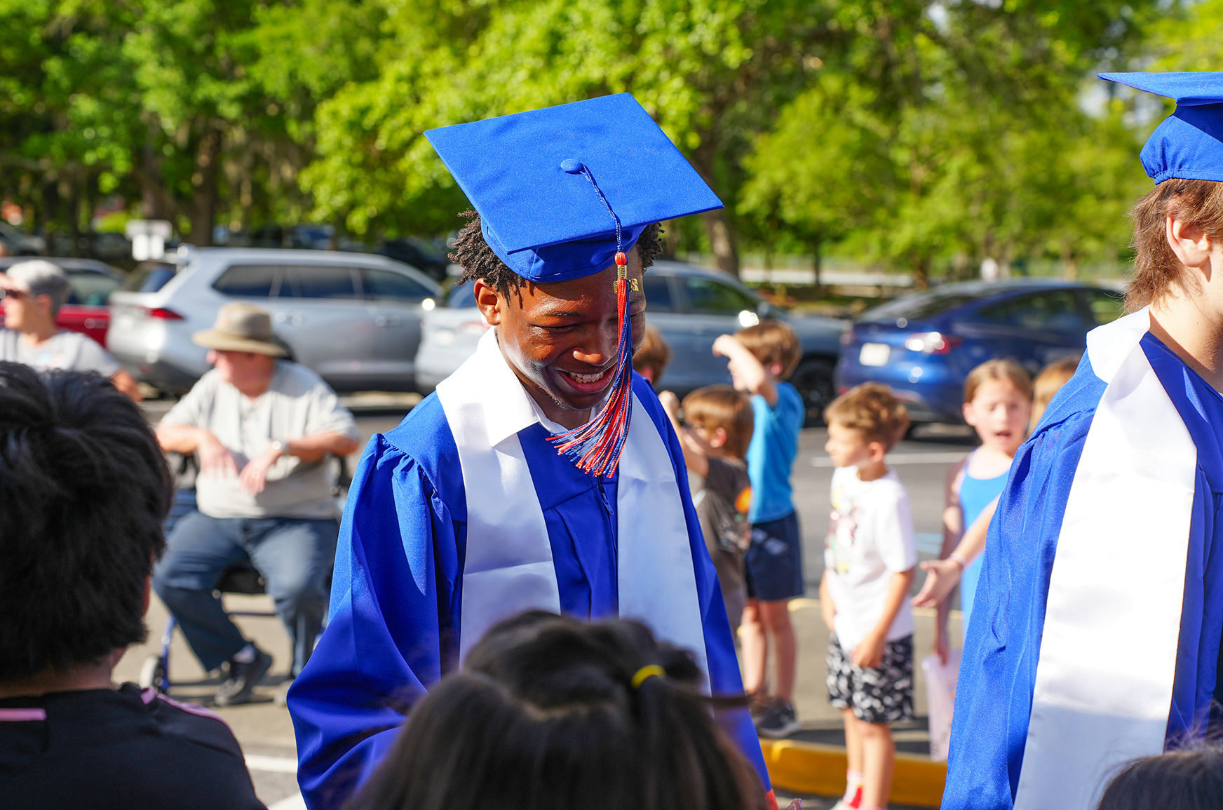 A person in a blue graduation cap and gown smiles in a parking lot with others.