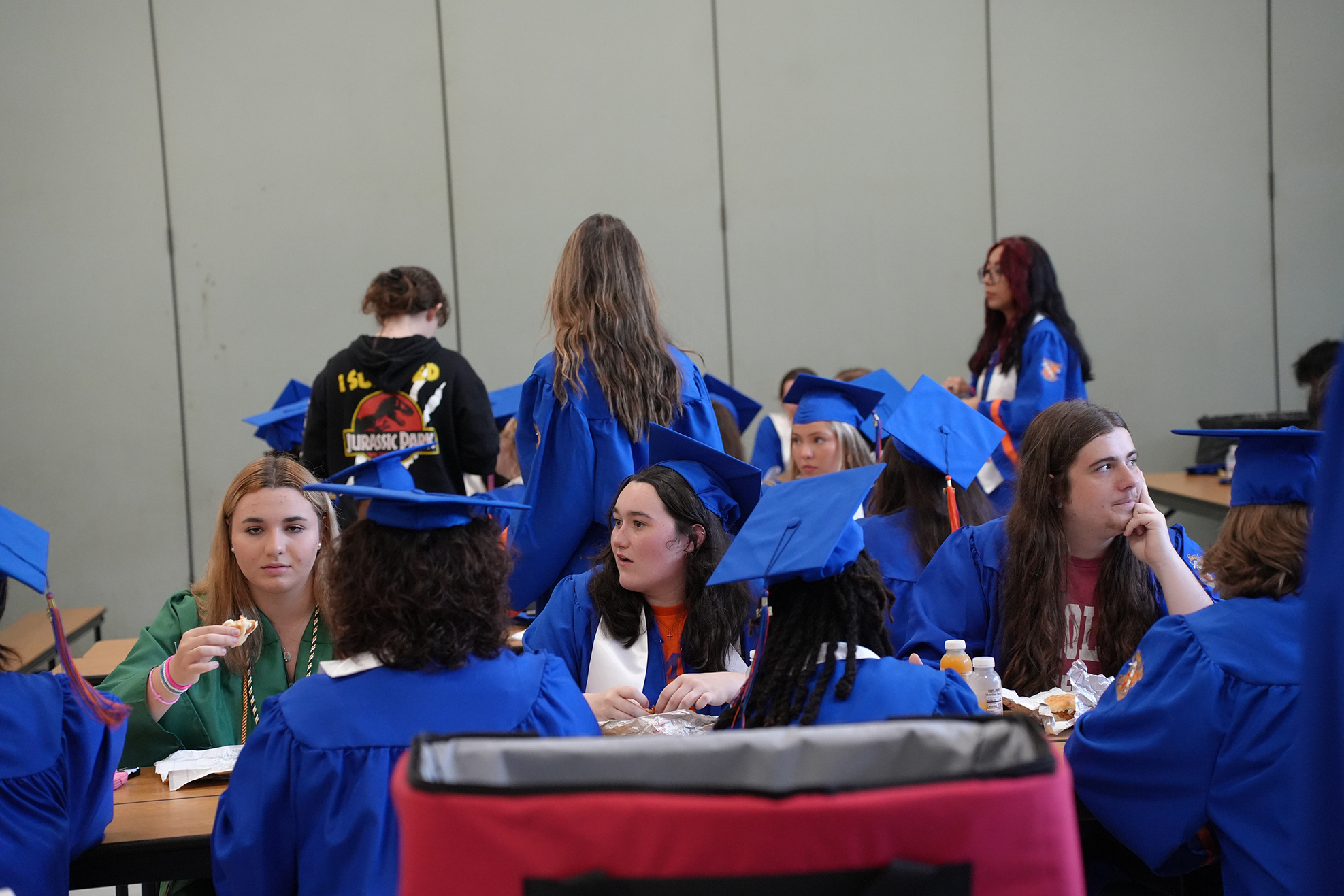 Graduates in blue gowns and hats seated at a table in a room, some eating and some talking.
