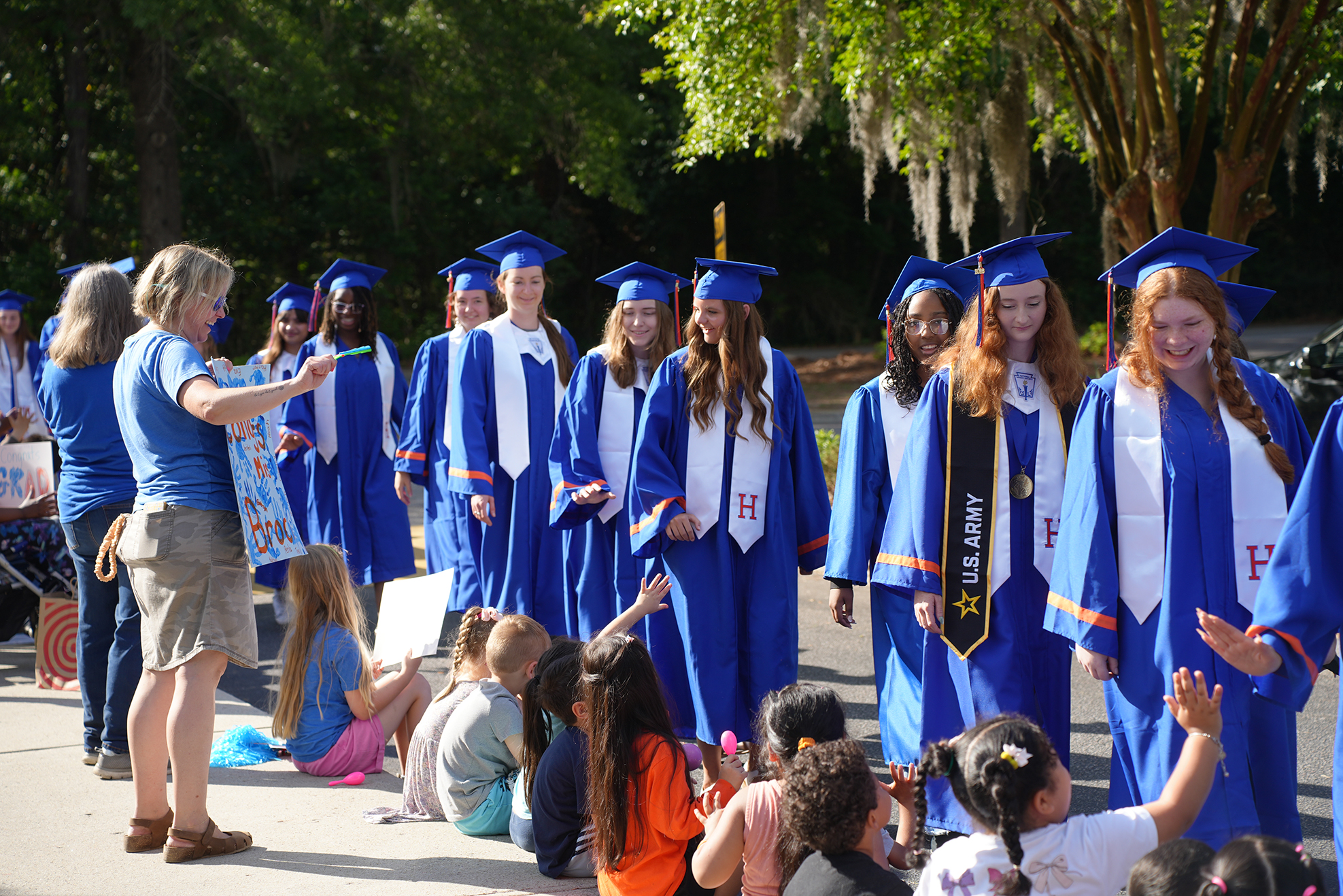 People in blue graduation gowns line up outdoors. A woman in blue stands and talks. Children sit in front of them.