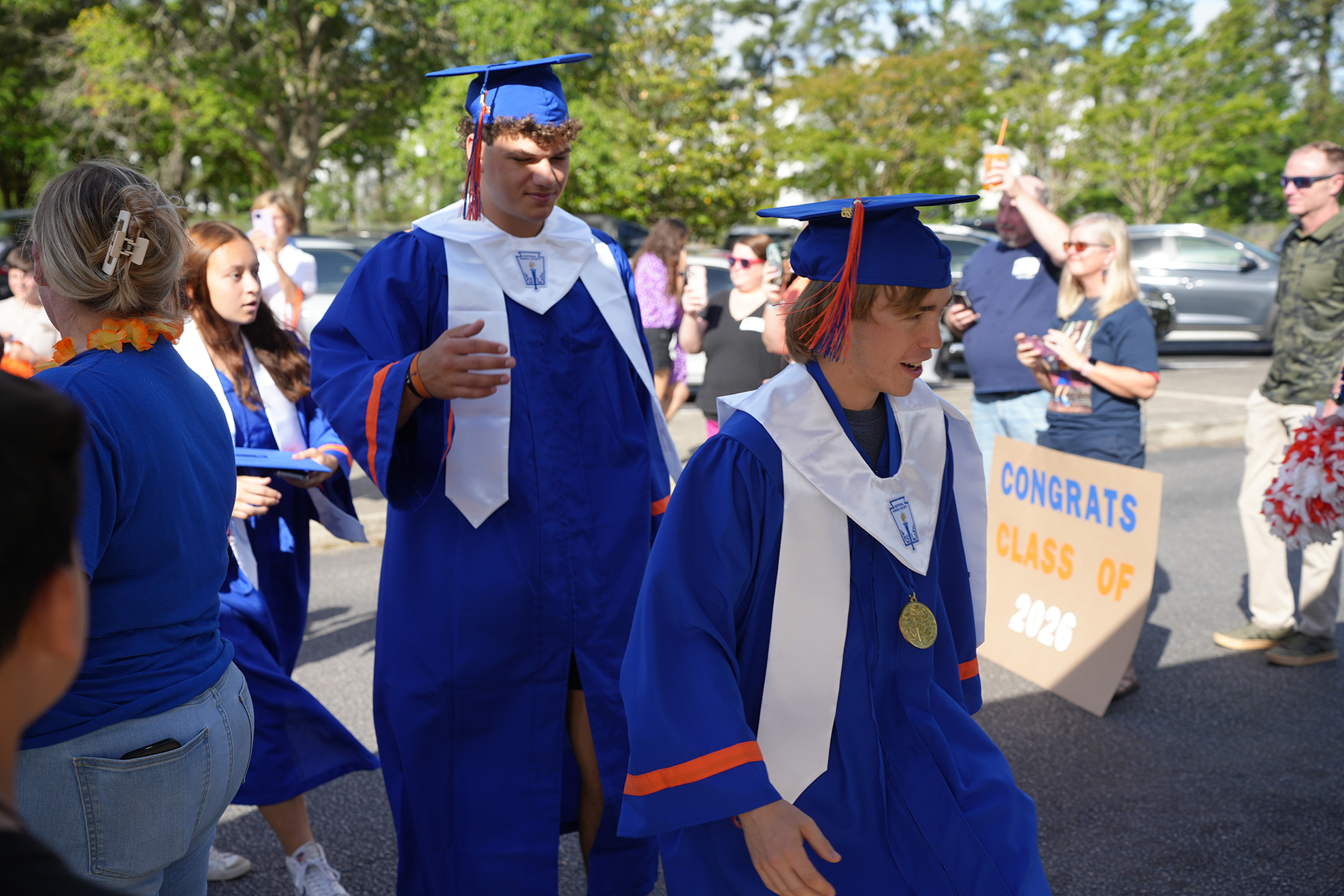 A group of graduates in blue robes and hats walk in a parade with others cheering.