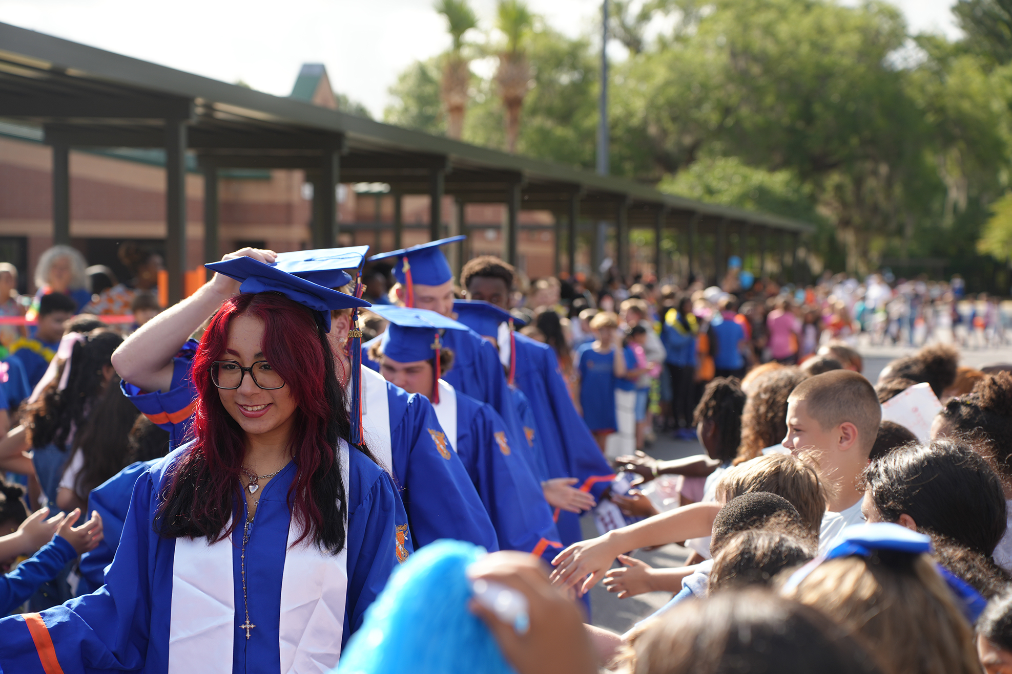 Graduates in blue caps and gowns walk through a crowd, under covered walkways, in front of a building.