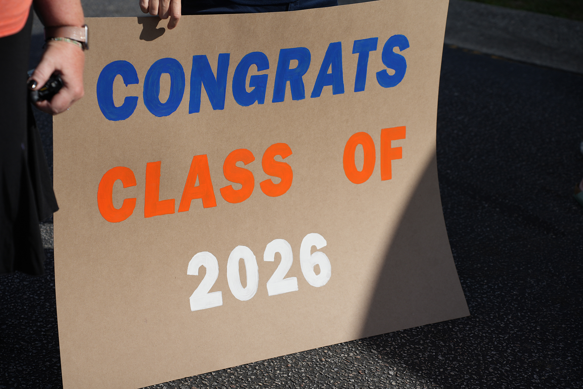 A person holds a cardboard sign with the words "CONGRATS CLASS OF 2026" written in blue, orange, and white fonts.
