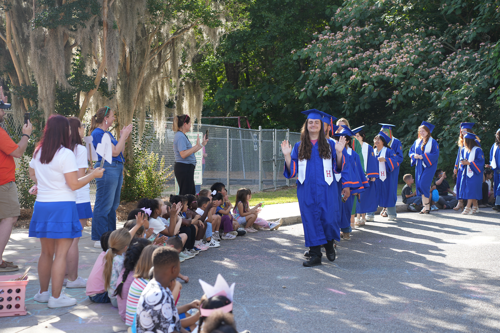 Graduates in blue gowns walk down a road, while others watch from a sidewalk. Some children sit on the ground.