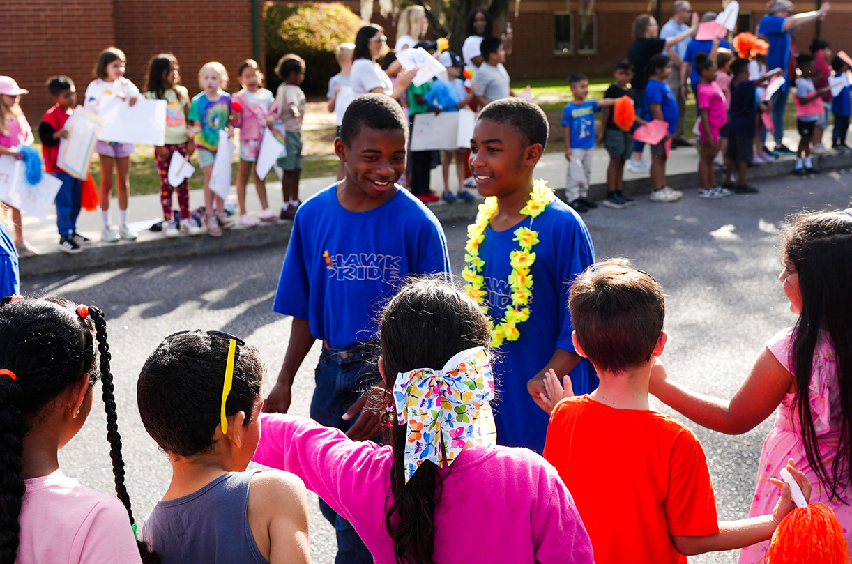 Children gather in a circle; two boys wear blue shirts, one with a yellow garland. Other children stand behind them, some holding signs.