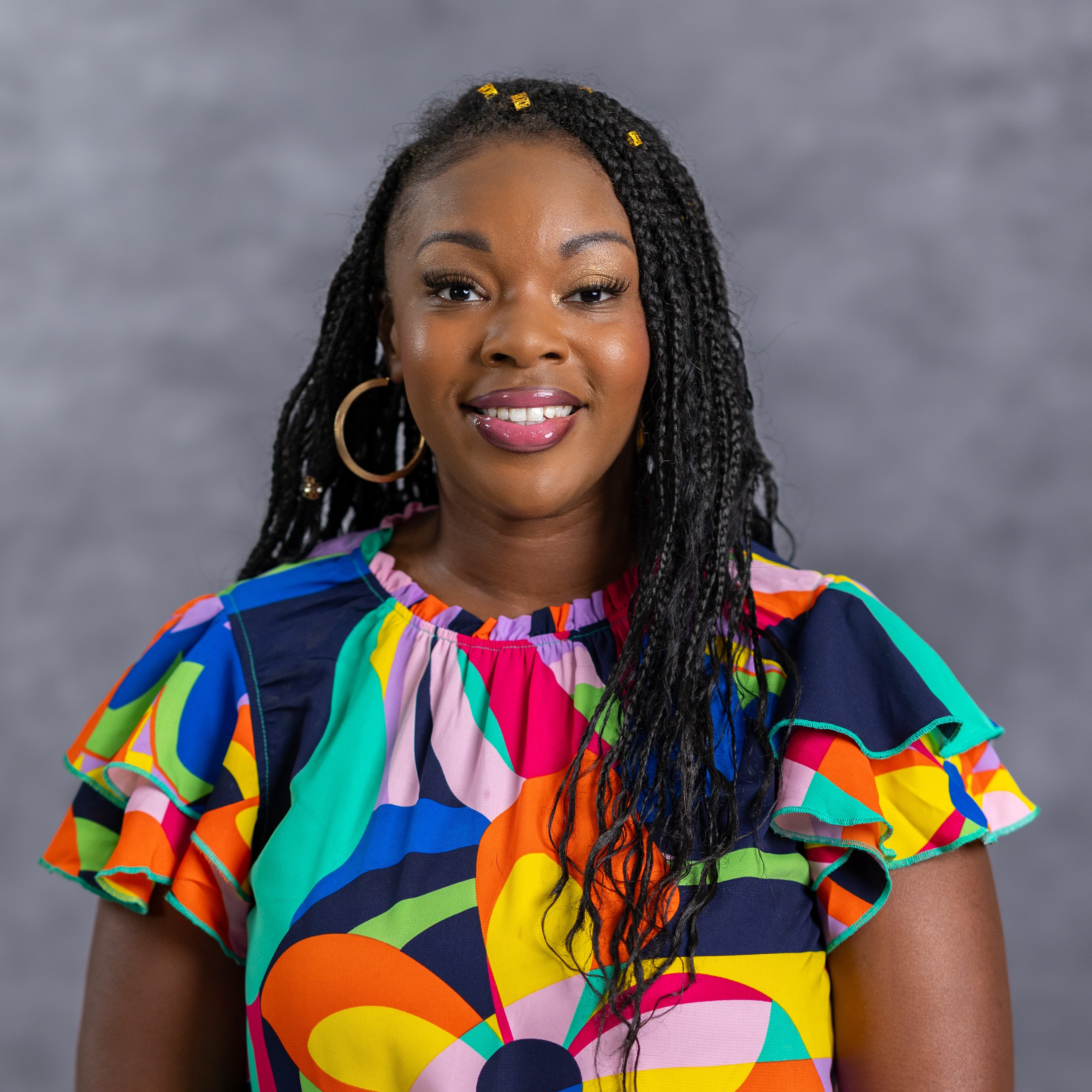 Woman with braids, wearing a colorful blouse with a flower design, standing against a gray background.