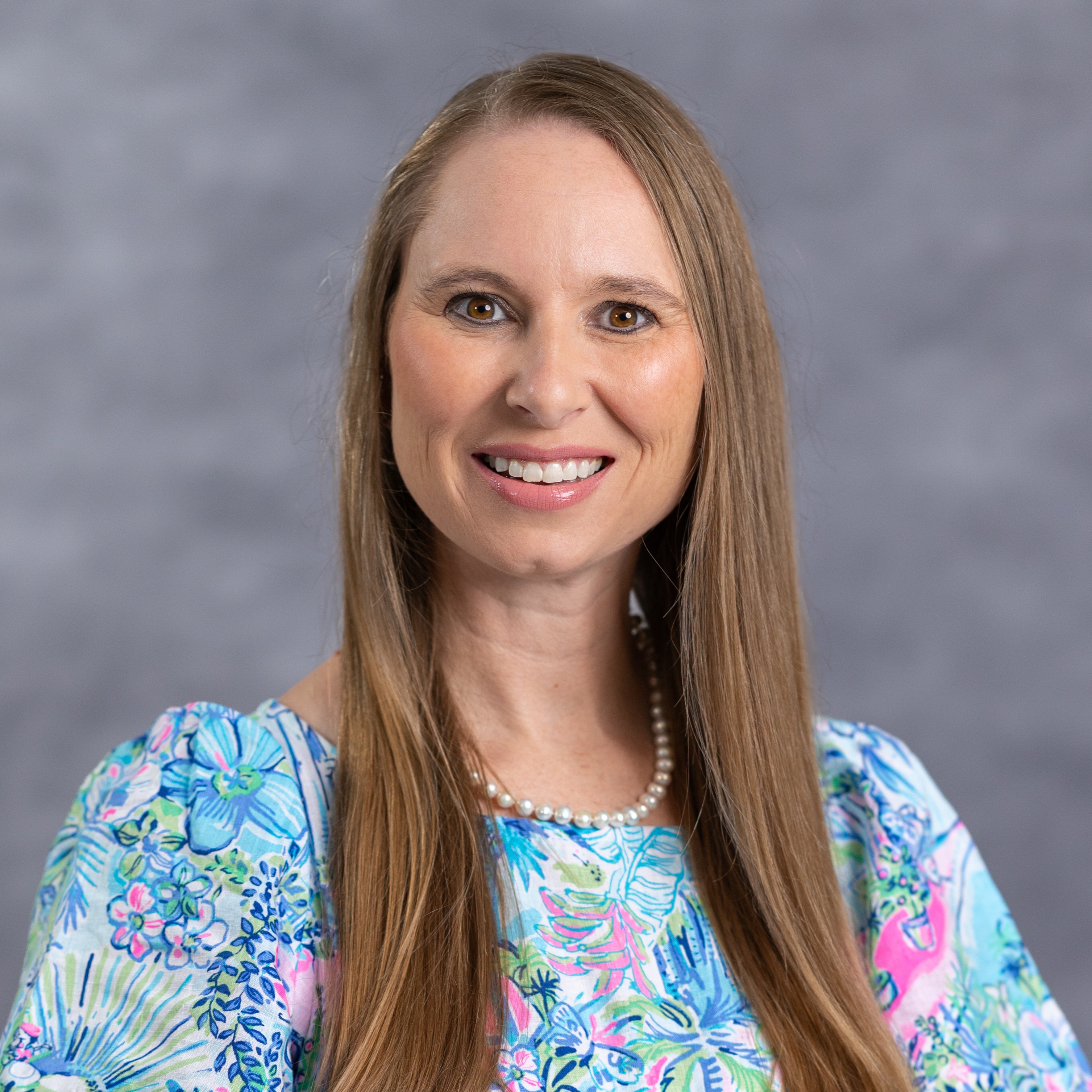 Woman with brown hair in a floral dress and pearl necklace smiles against a gray background.