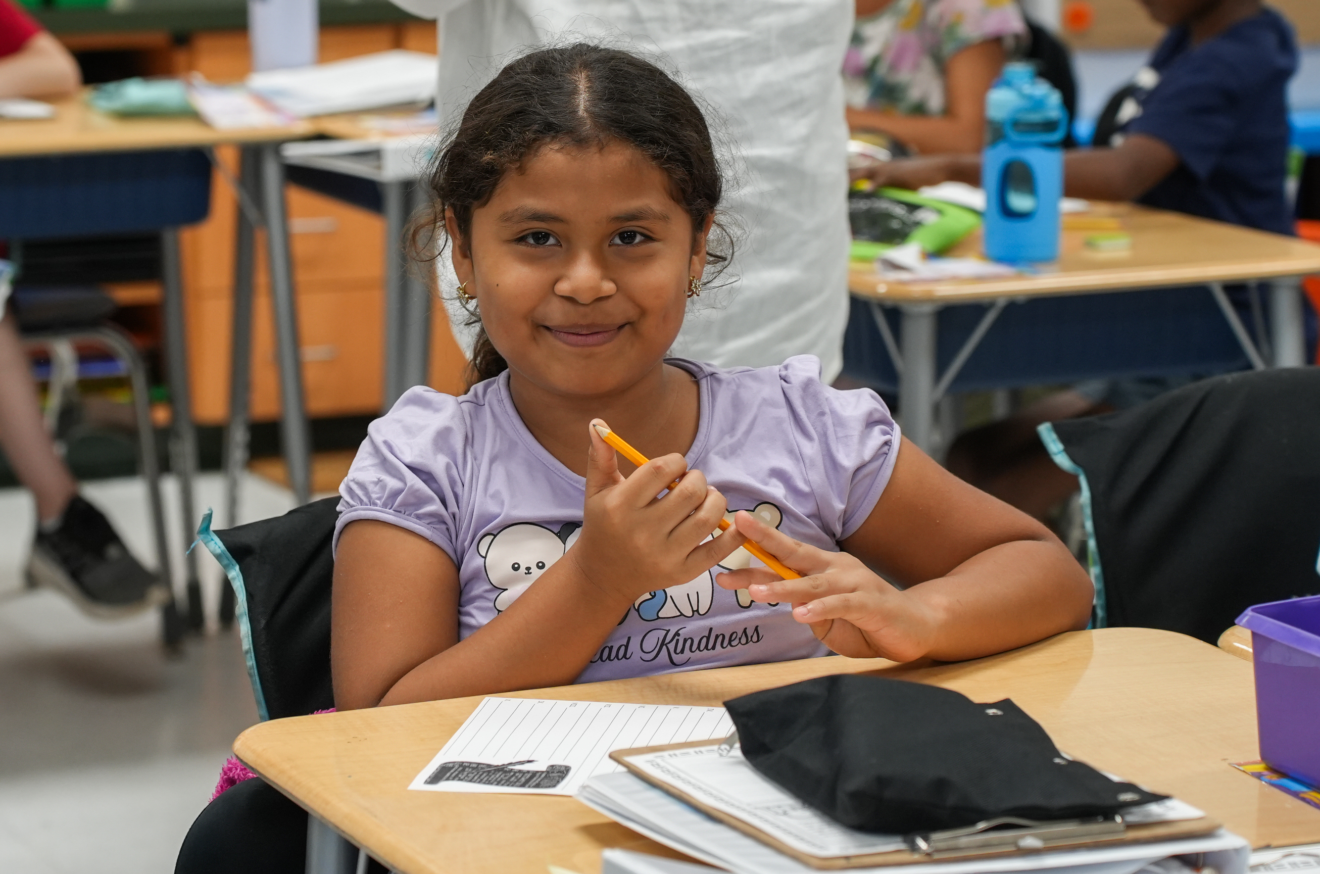A young girl in a classroom setting, holding a pencil and smiling, sits at a desk with papers and a black bag.