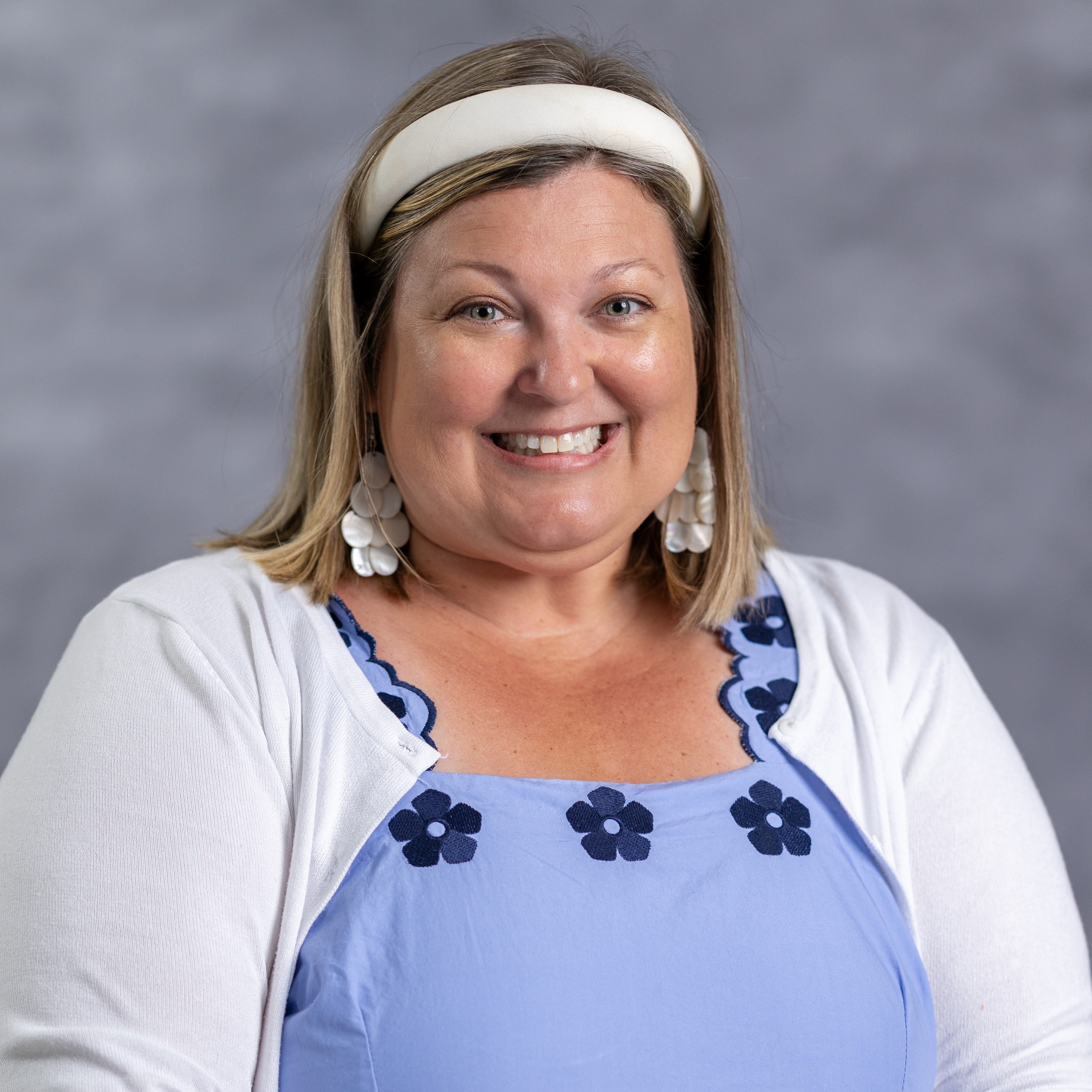 A woman with blonde hair smiles, wearing a light blue blouse with white flowers, a white cardigan, and a white headband.