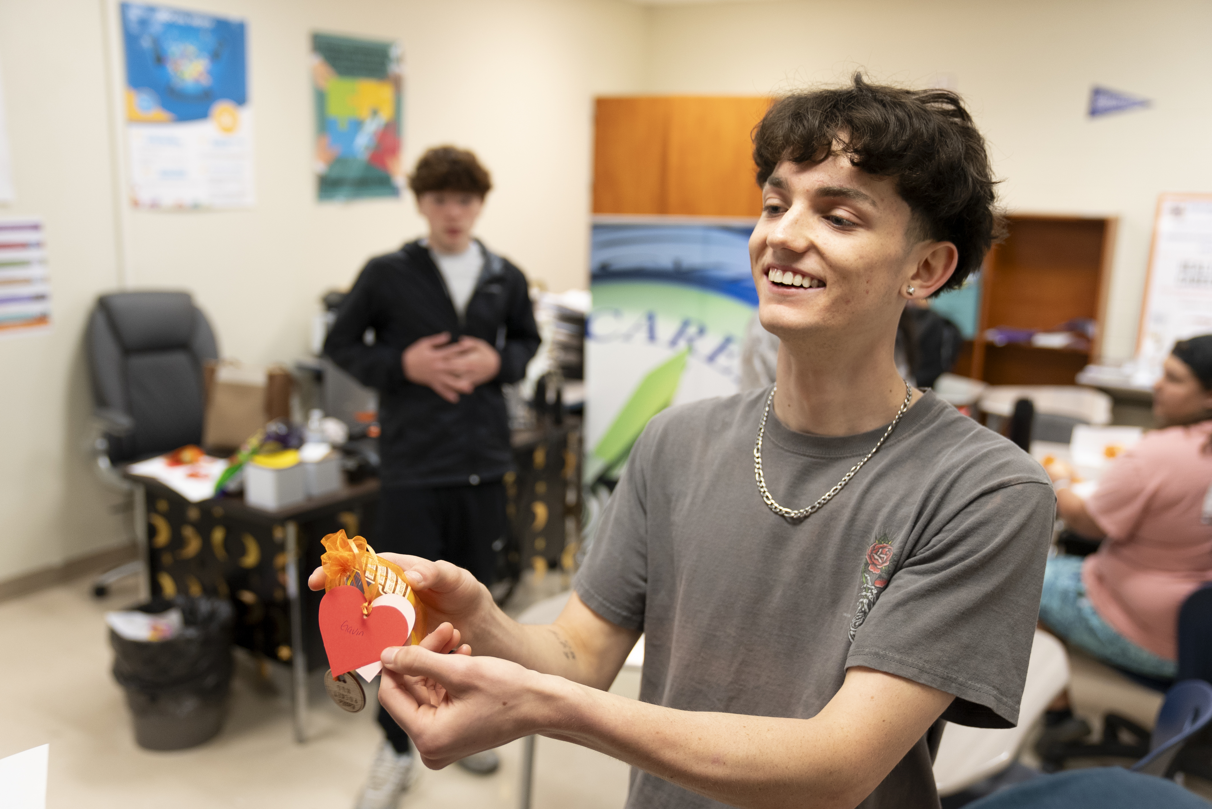 A boy in a gray T-shirt holds up a heart made of paper while smiling. Another student stands behind him.