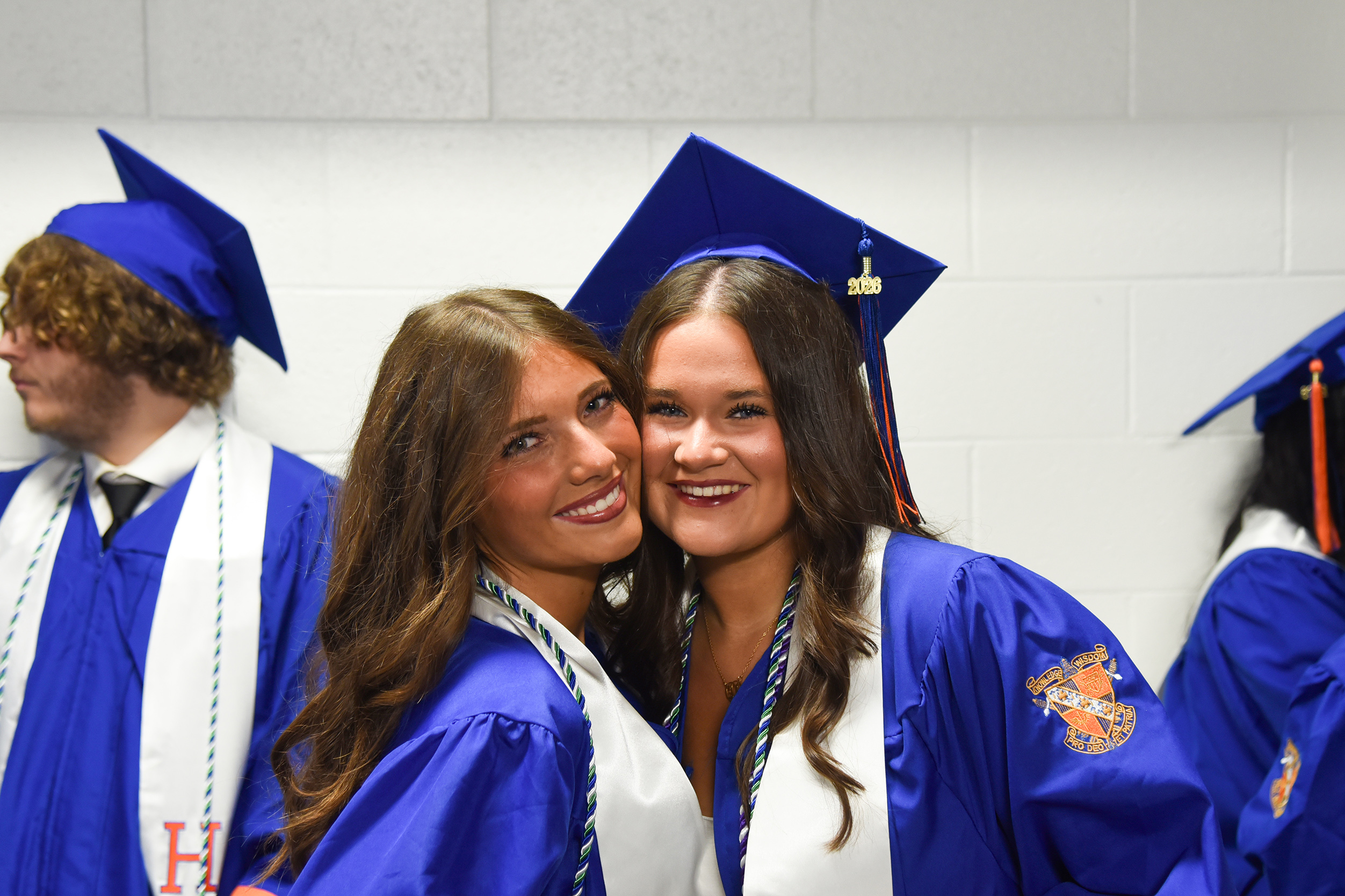 Two women in blue graduation gowns pose for a photo. Their caps are tilted and they smile.