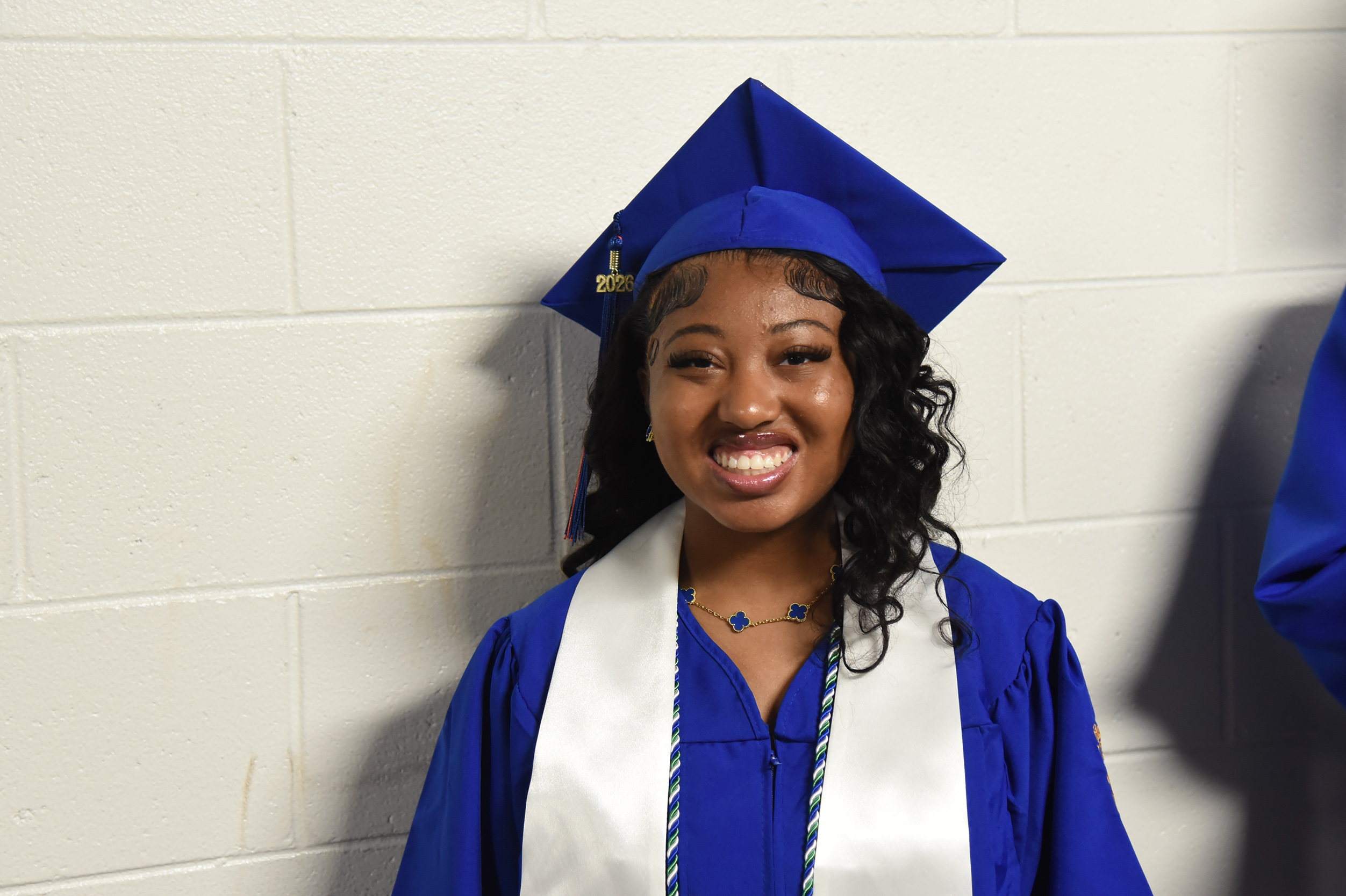 A smiling woman in a blue graduation gown and hat stands against a white brick wall.