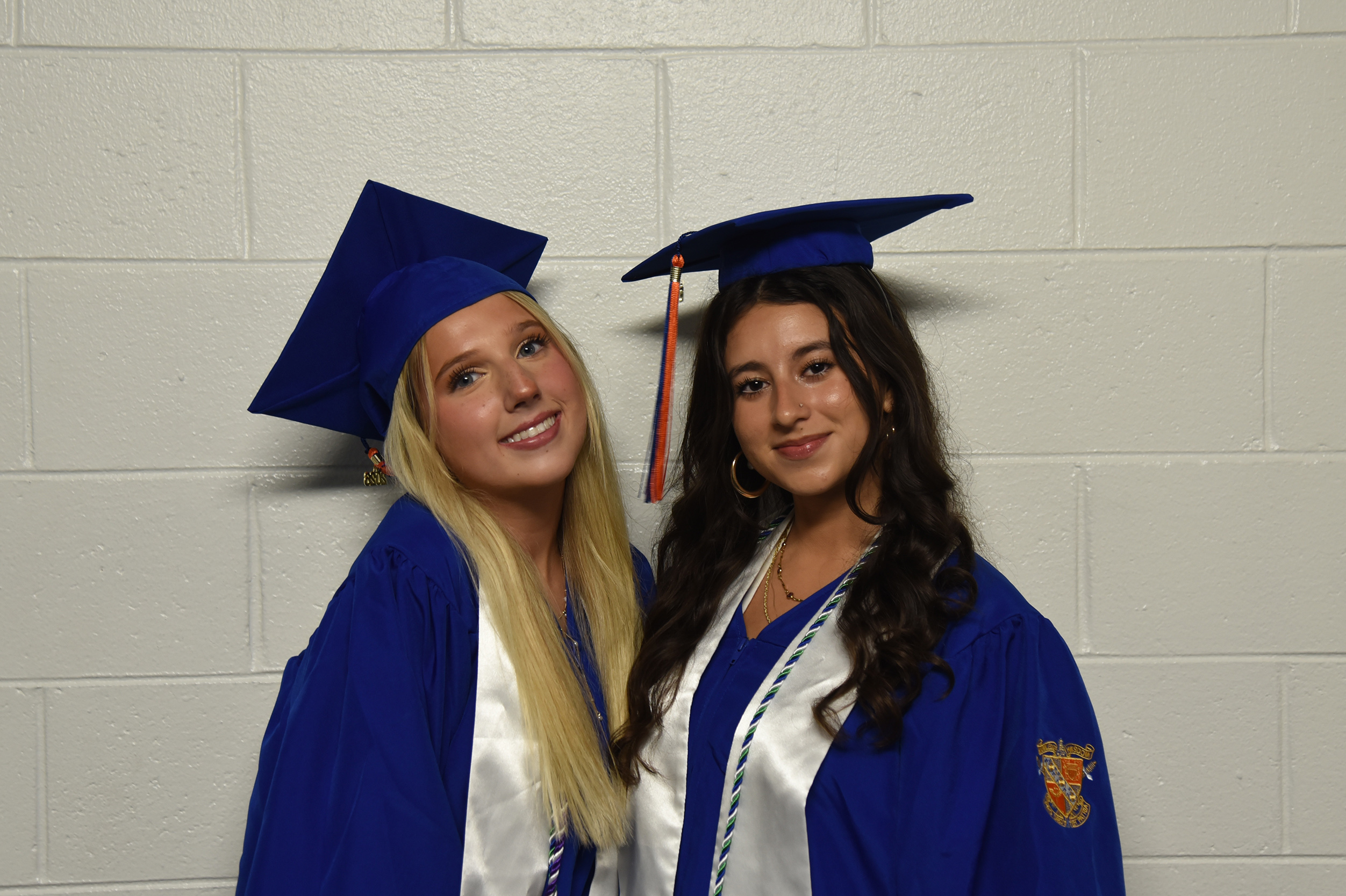 Two women in blue graduation gowns and caps stand together. One wears a blue graduation tassel.