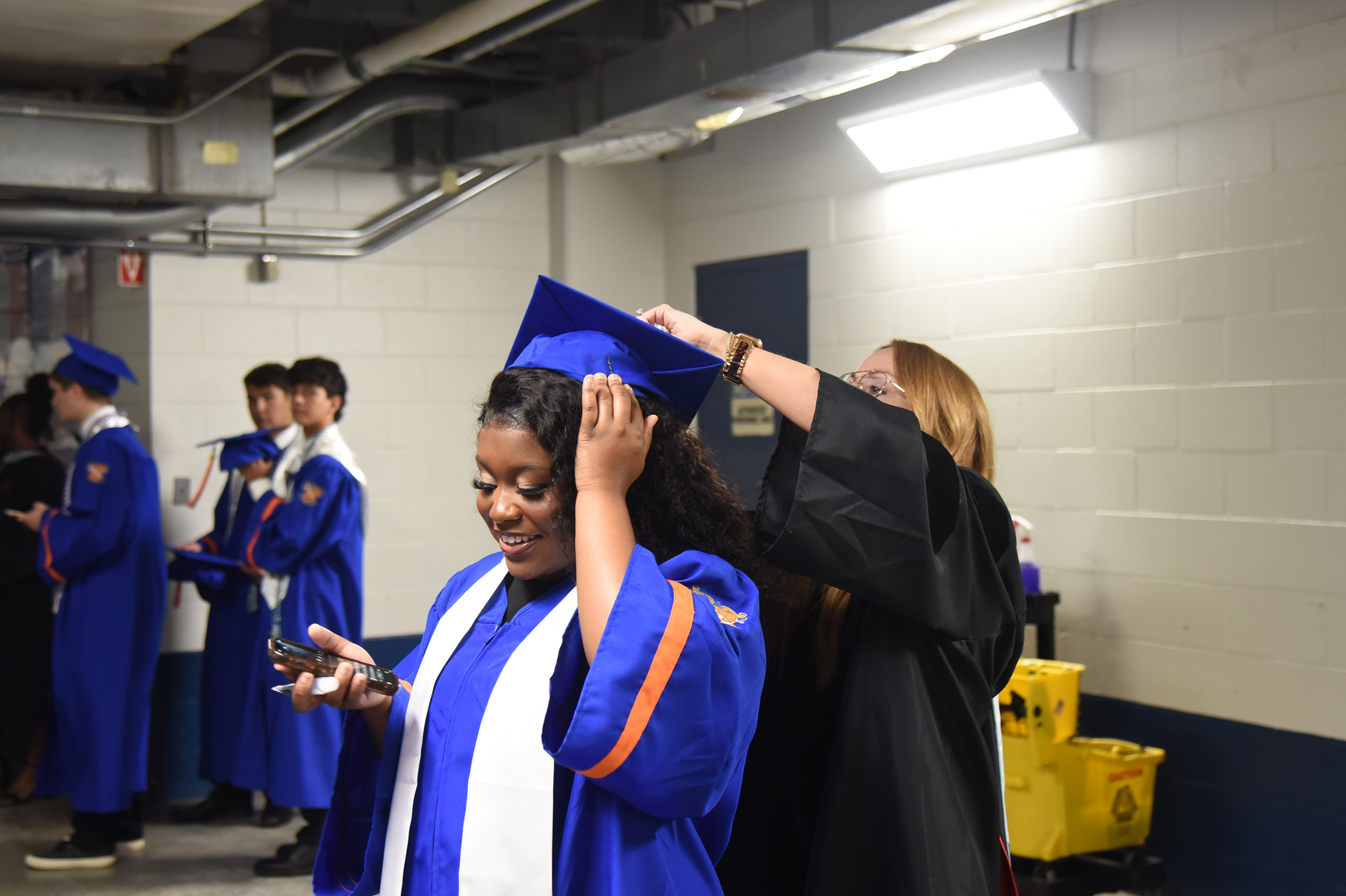 Graduates in blue gowns, one adjusting a cap, with a woman in black, white walls, and a yellow bin.