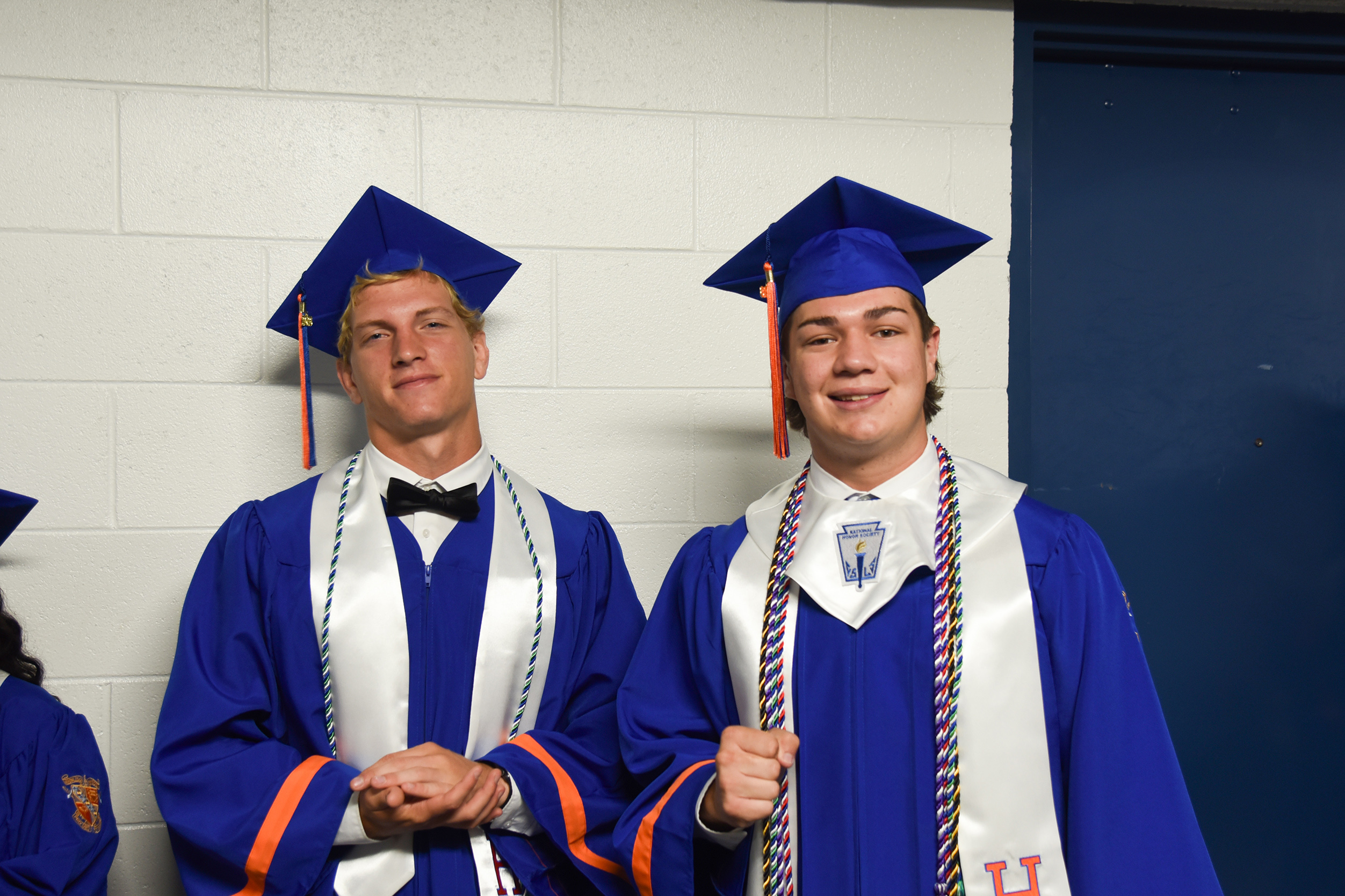 Two men in blue graduation gowns and caps pose, one wearing a bow tie and the other a sash.