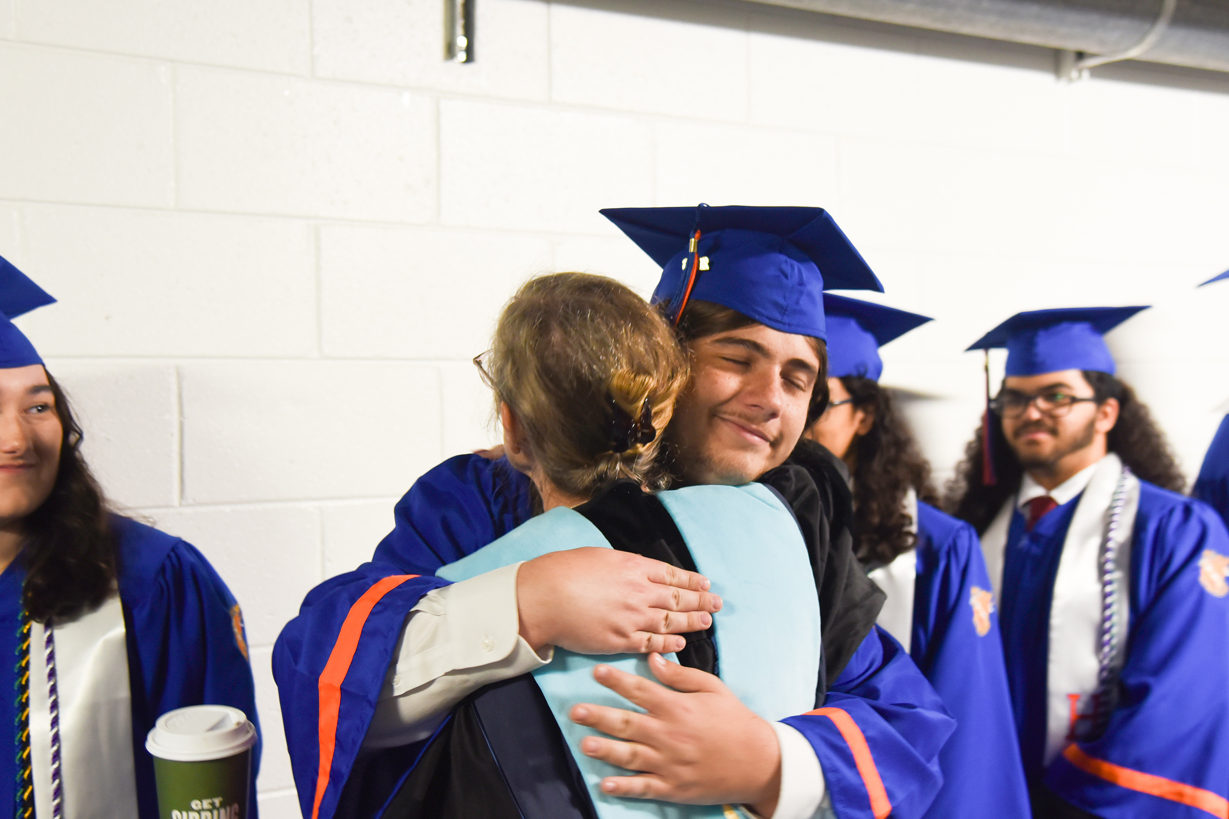 Graduates in blue and white gowns and caps, one couple embracing, others standing near a wall.