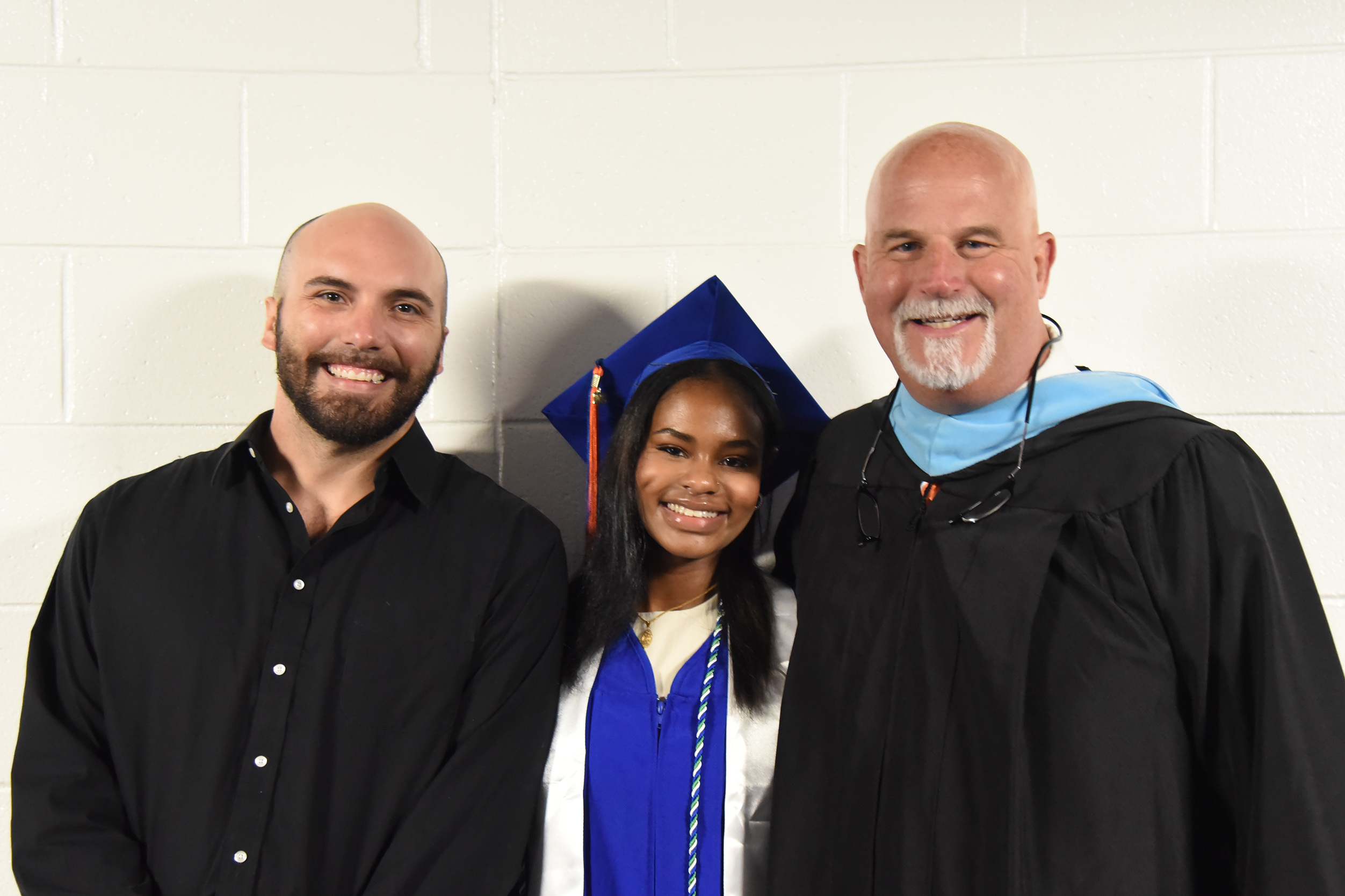 Three people smile at the camera. A young woman wears a blue graduation cap. Two men wear black attire.