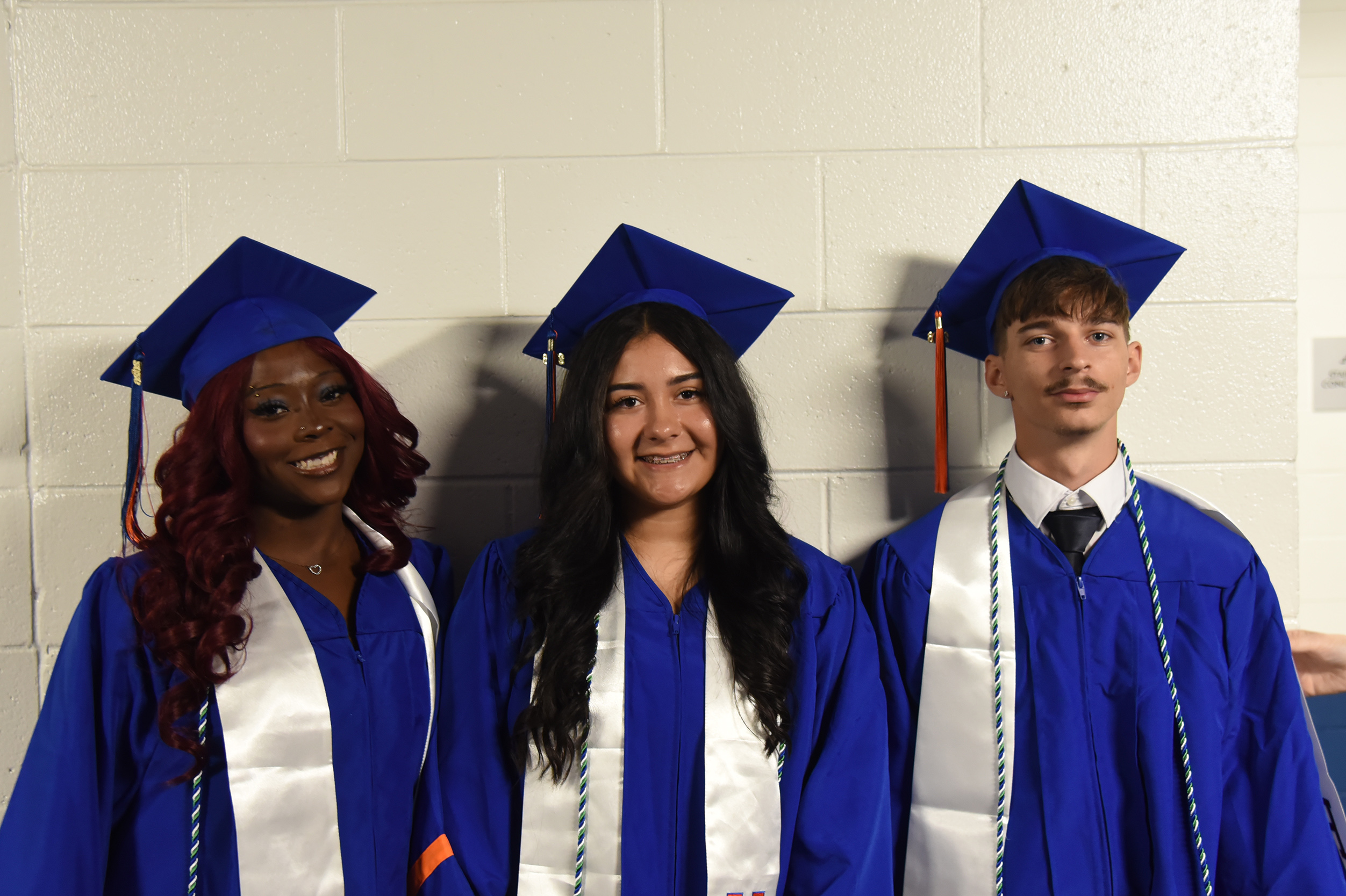 Three graduates in blue caps and gowns smile, standing against a white brick wall.