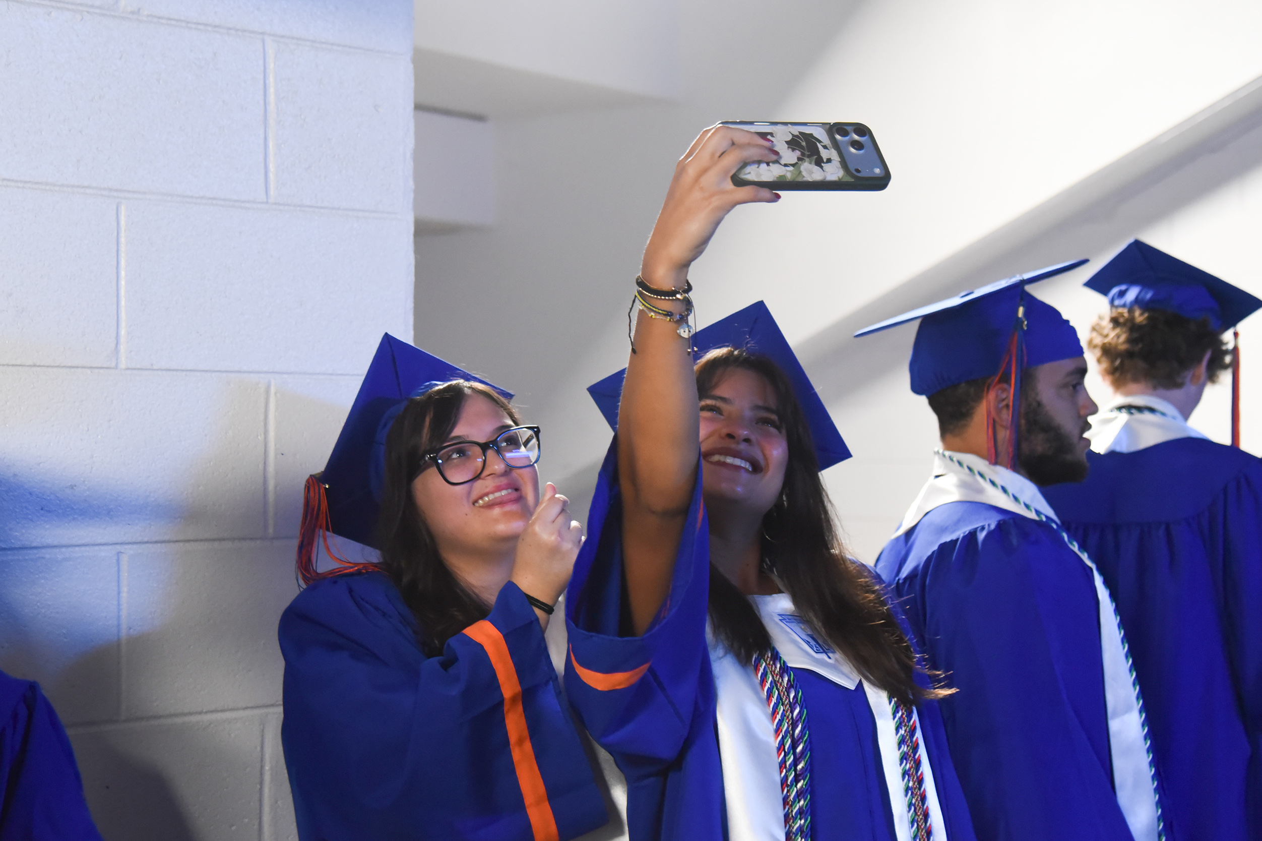 People in blue graduation gowns take a selfie with their phones; one wears glasses.