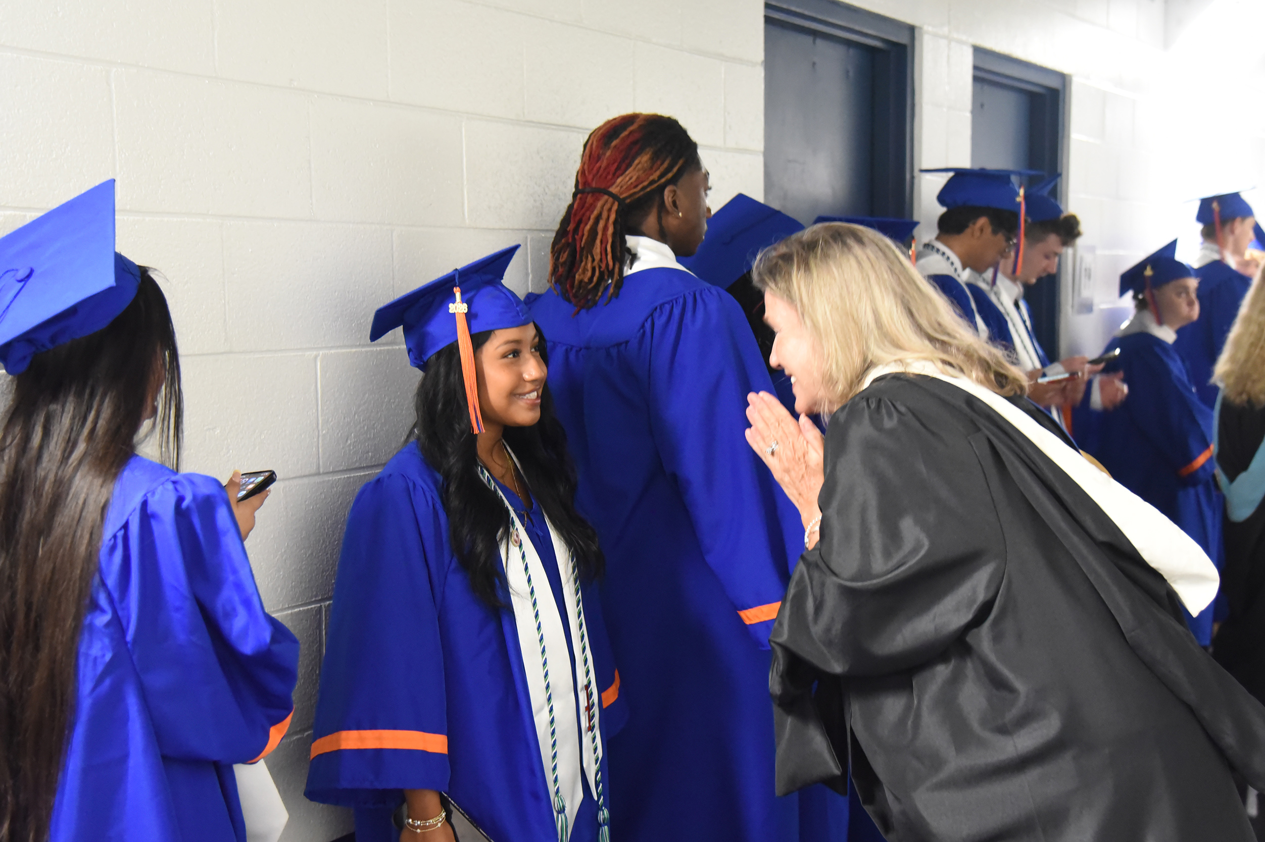 Graduates in blue gowns and caps stand in line; a woman in a black gown talks to a smiling graduate.