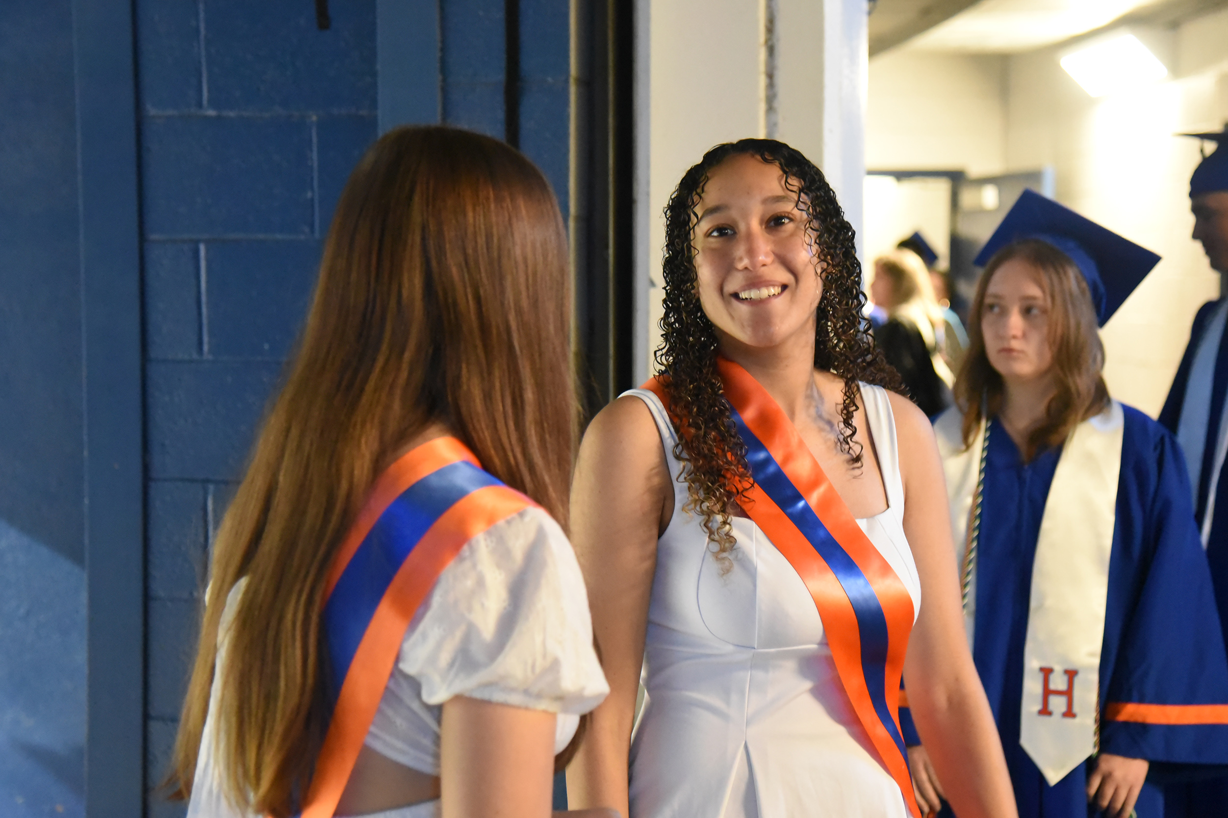 Three women in caps and gowns stand close together. Two wear sashes. One woman smiles.