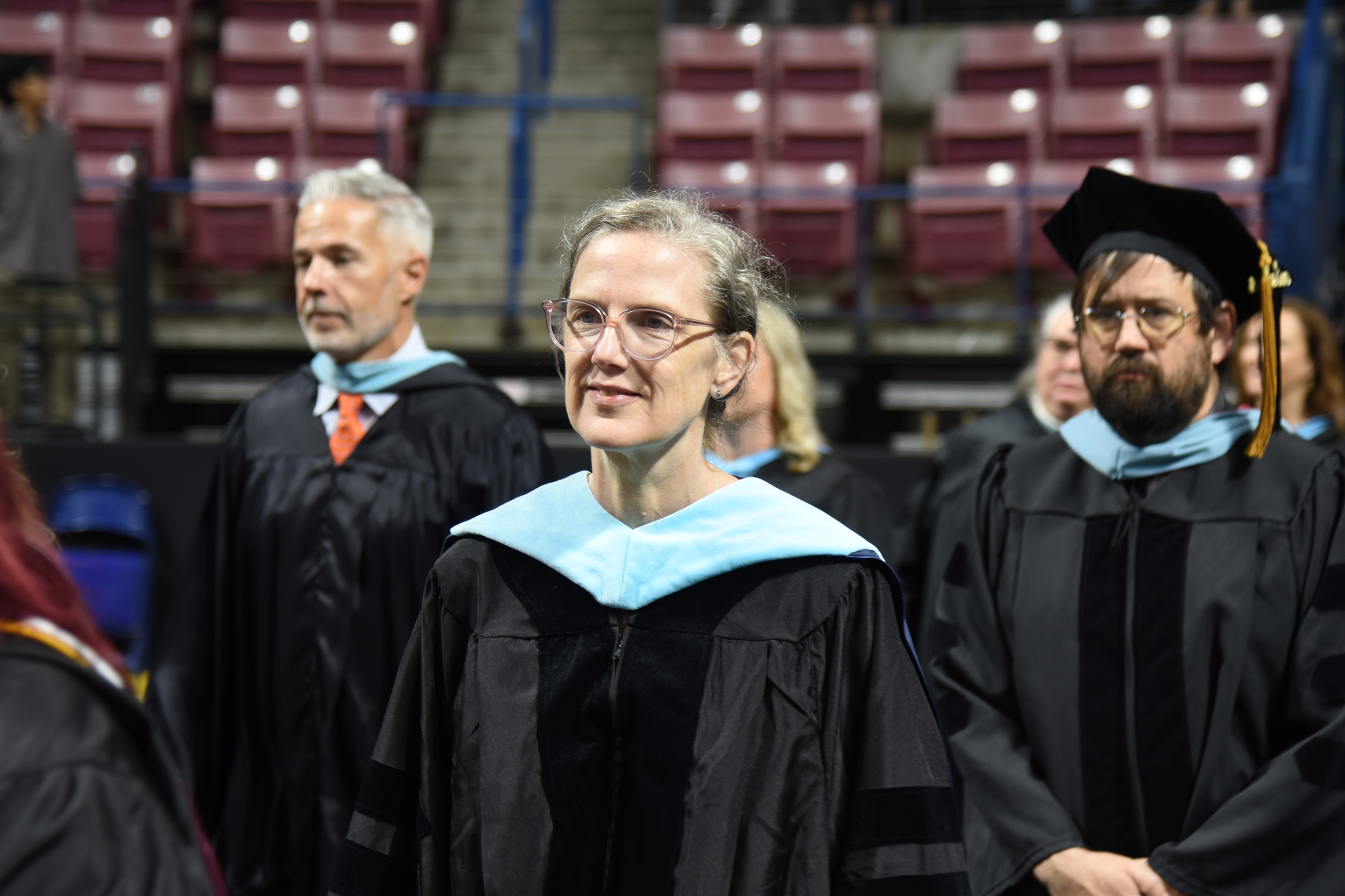Graduates in black gowns and caps in an arena. The woman in the front wears glasses and smiles.